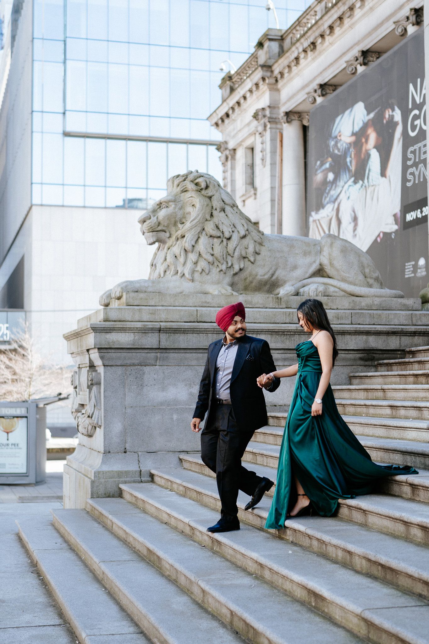 A couple in formal attire walks down grand stone steps beside an ornate lion statue at a historic building entrance.