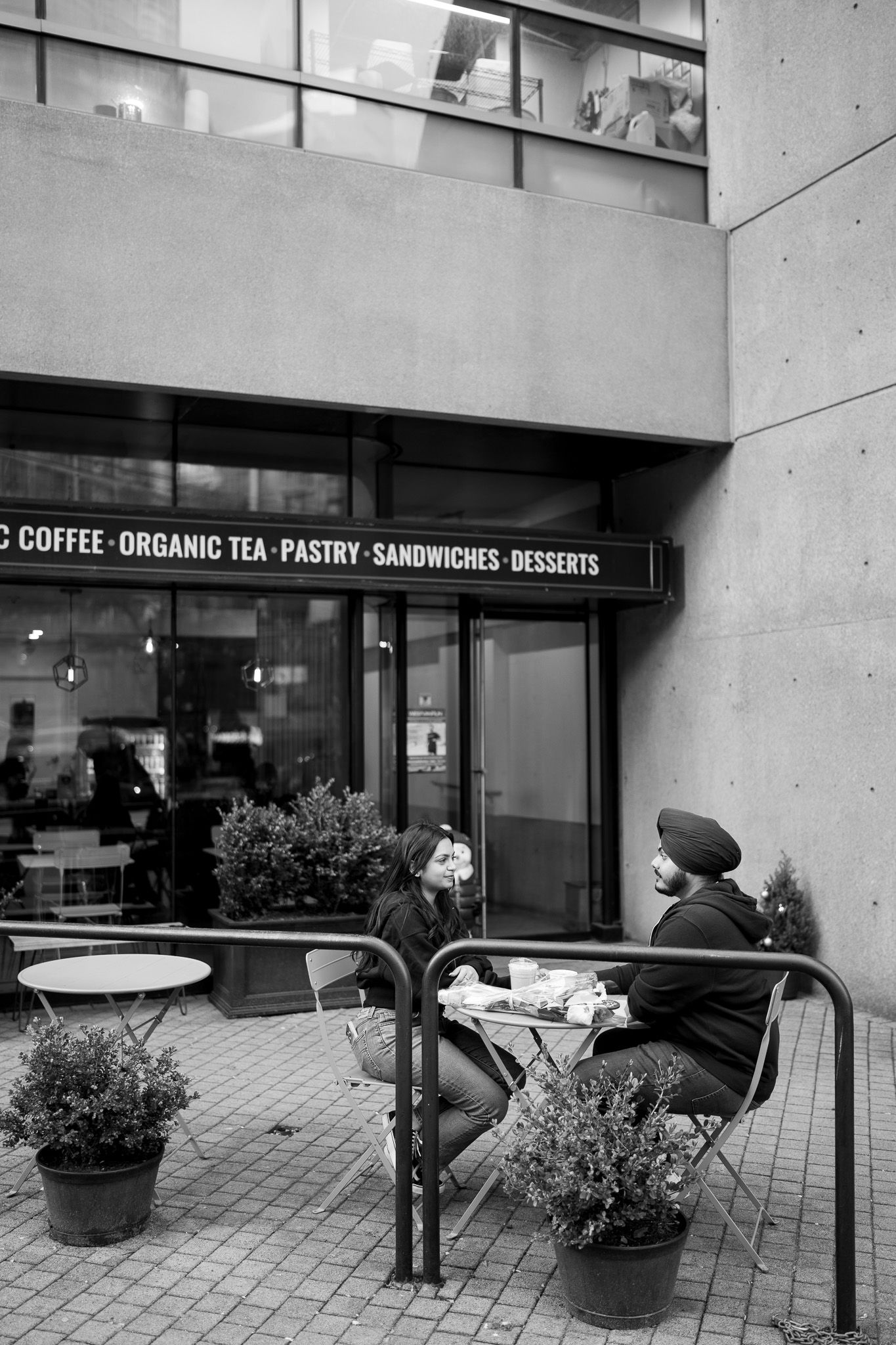 Modern cafe storefront with concrete walls, glass windows, and outdoor seating area featuring potted plants.