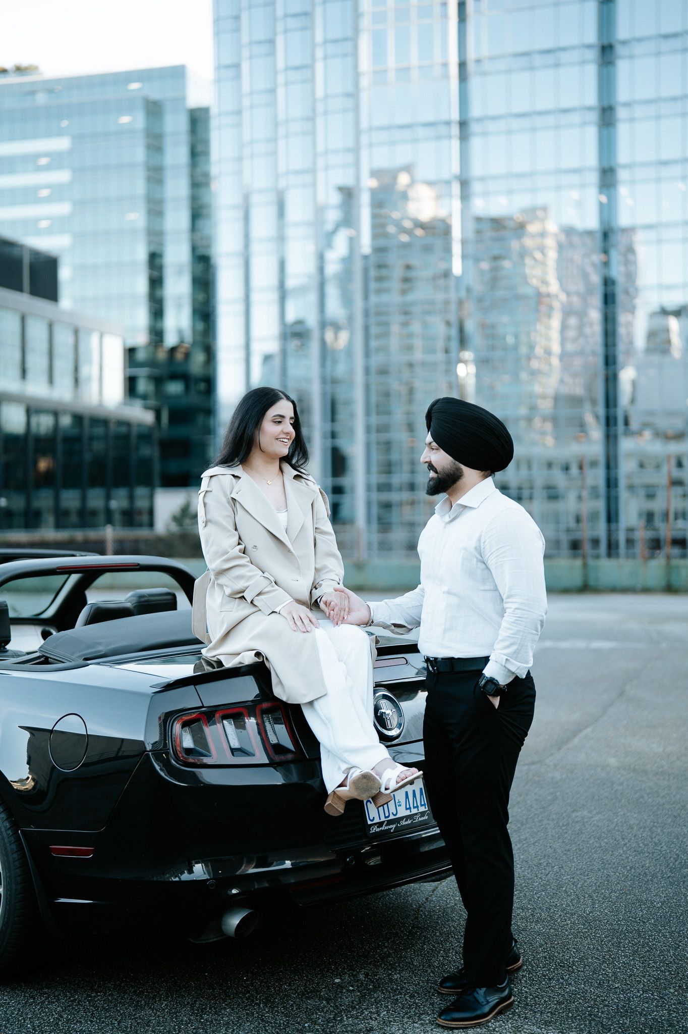 Two people having a conversation next to a black sports car in an urban setting with glass buildings in background.