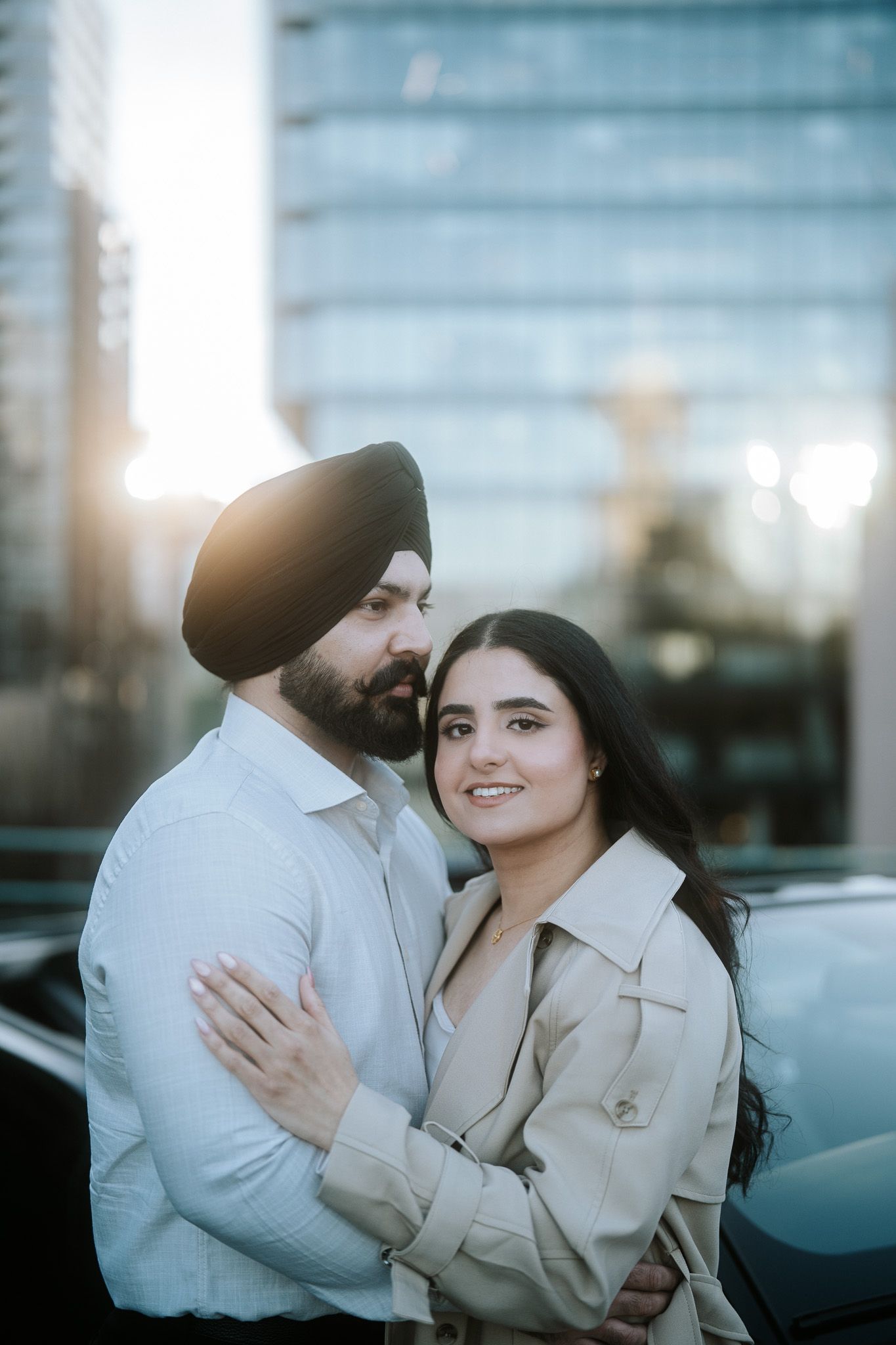 A romantic couple embraces outdoors in matching beige coats during golden hour in an urban setting.