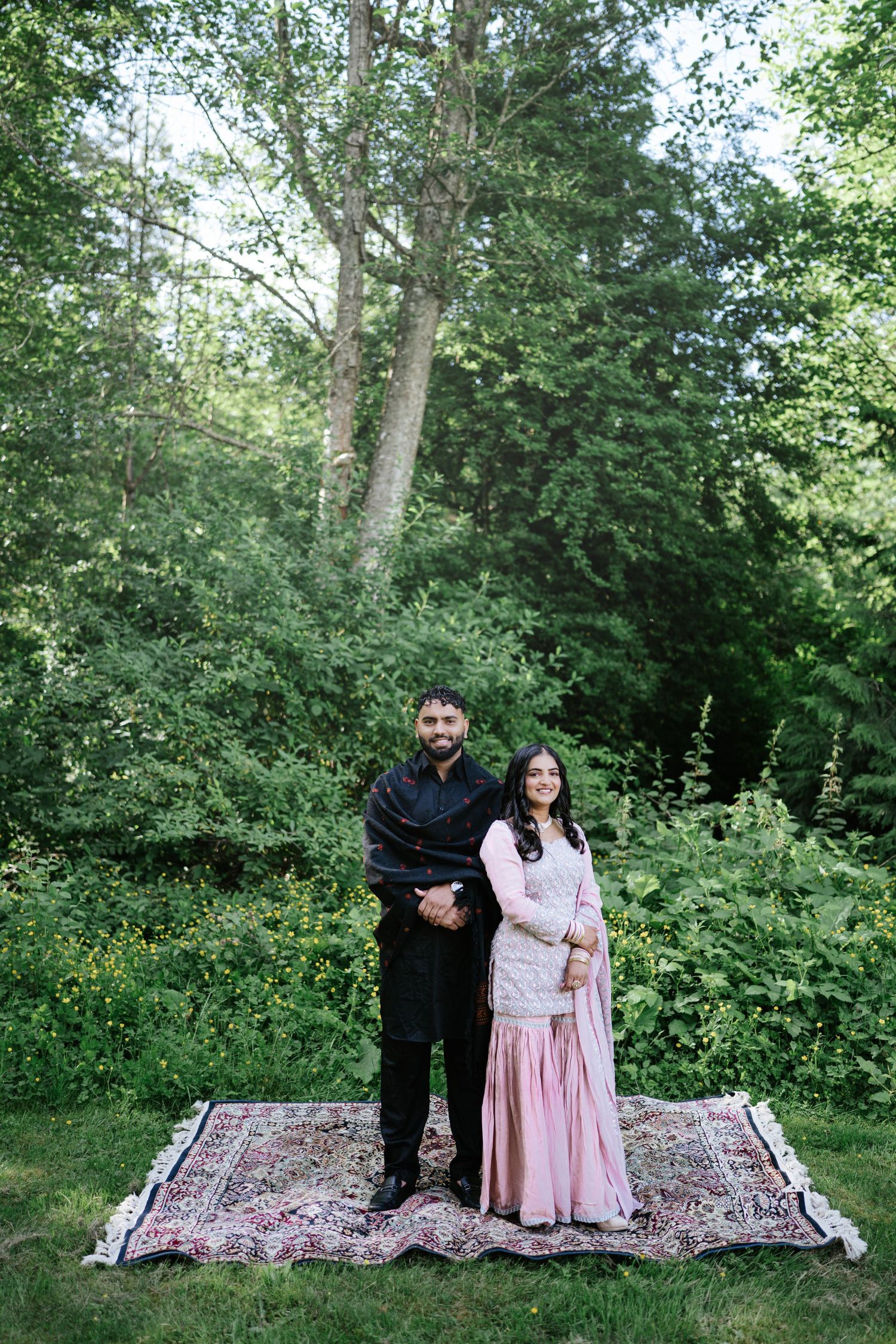 Couple poses together on decorative rugs laid out in a lush green forest setting.