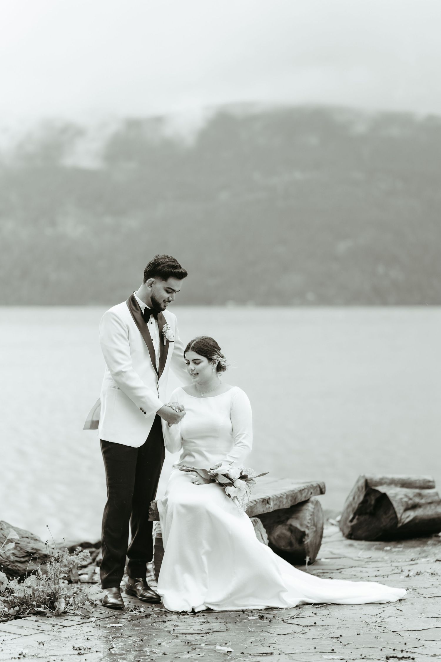 A couple in wedding attire sits together on a rocky beach shore in a vintage black and white photograph.