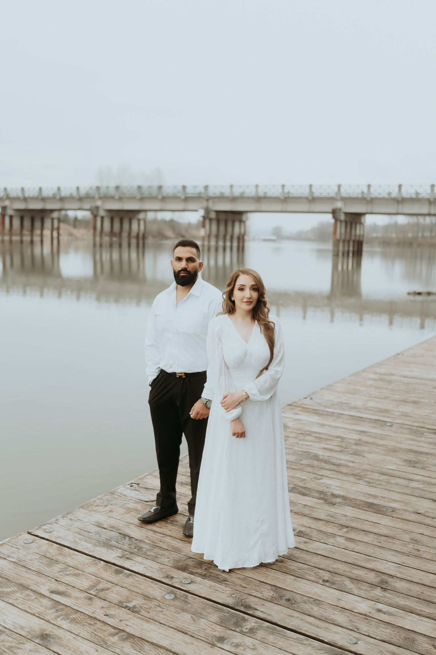 A couple in white and black formal wear poses on a wooden dock during an overcast day.
