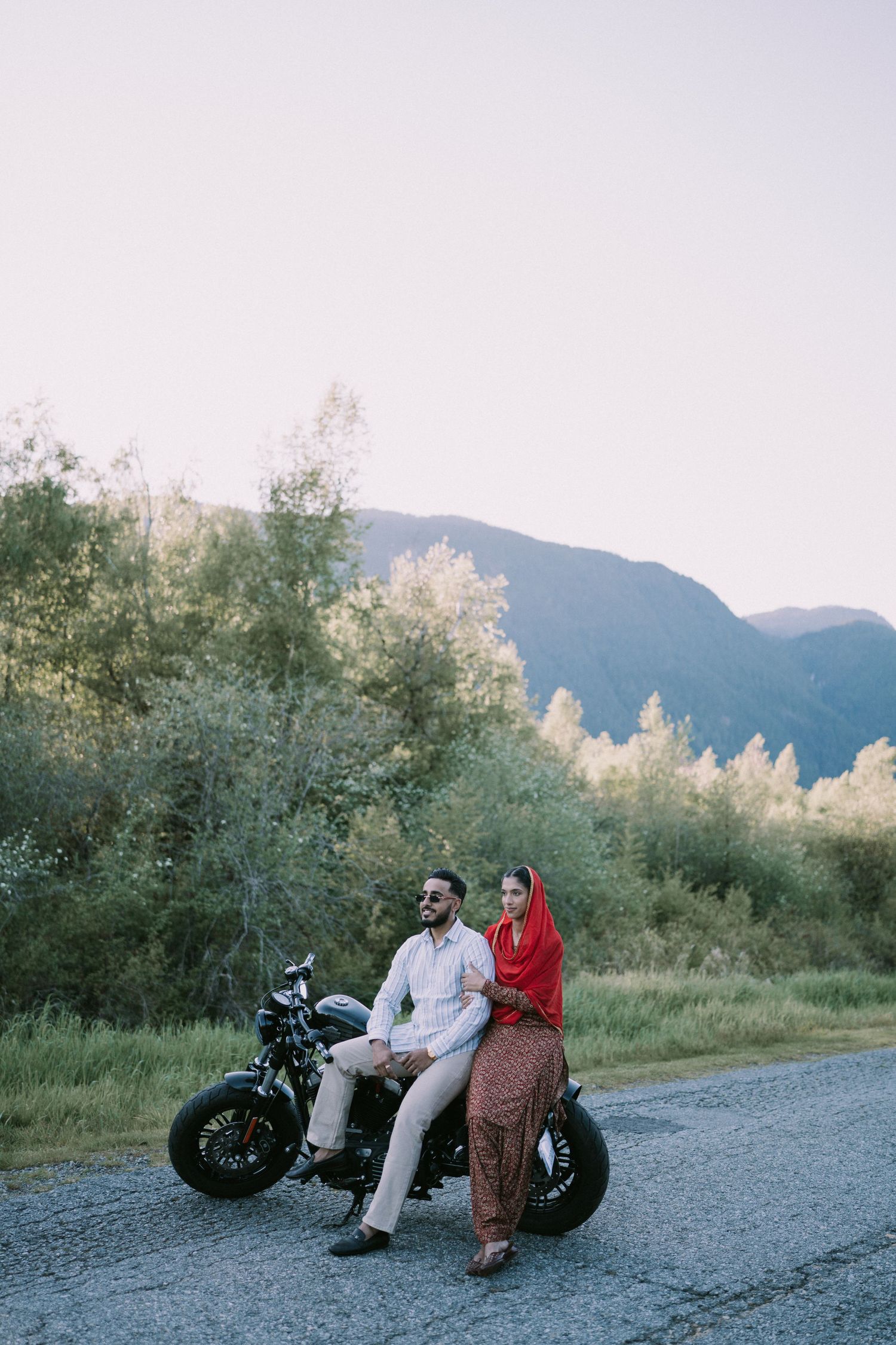 Two people sit together on a parked motorcycle against a scenic mountain backdrop at dusk.