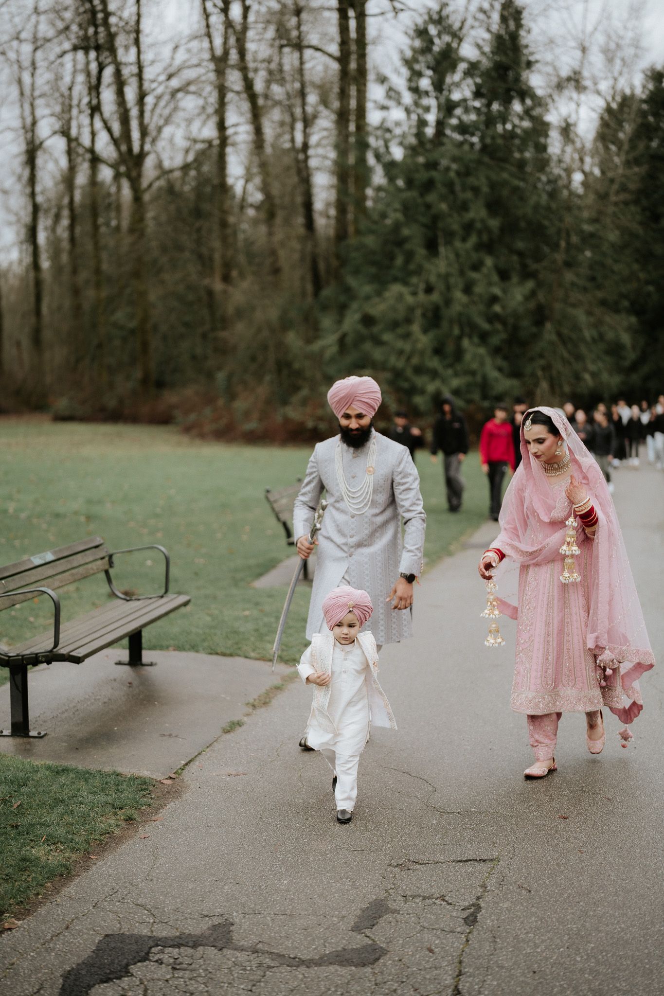A couple in pink and white outfits walking together in a park with benches and trees in the background.
