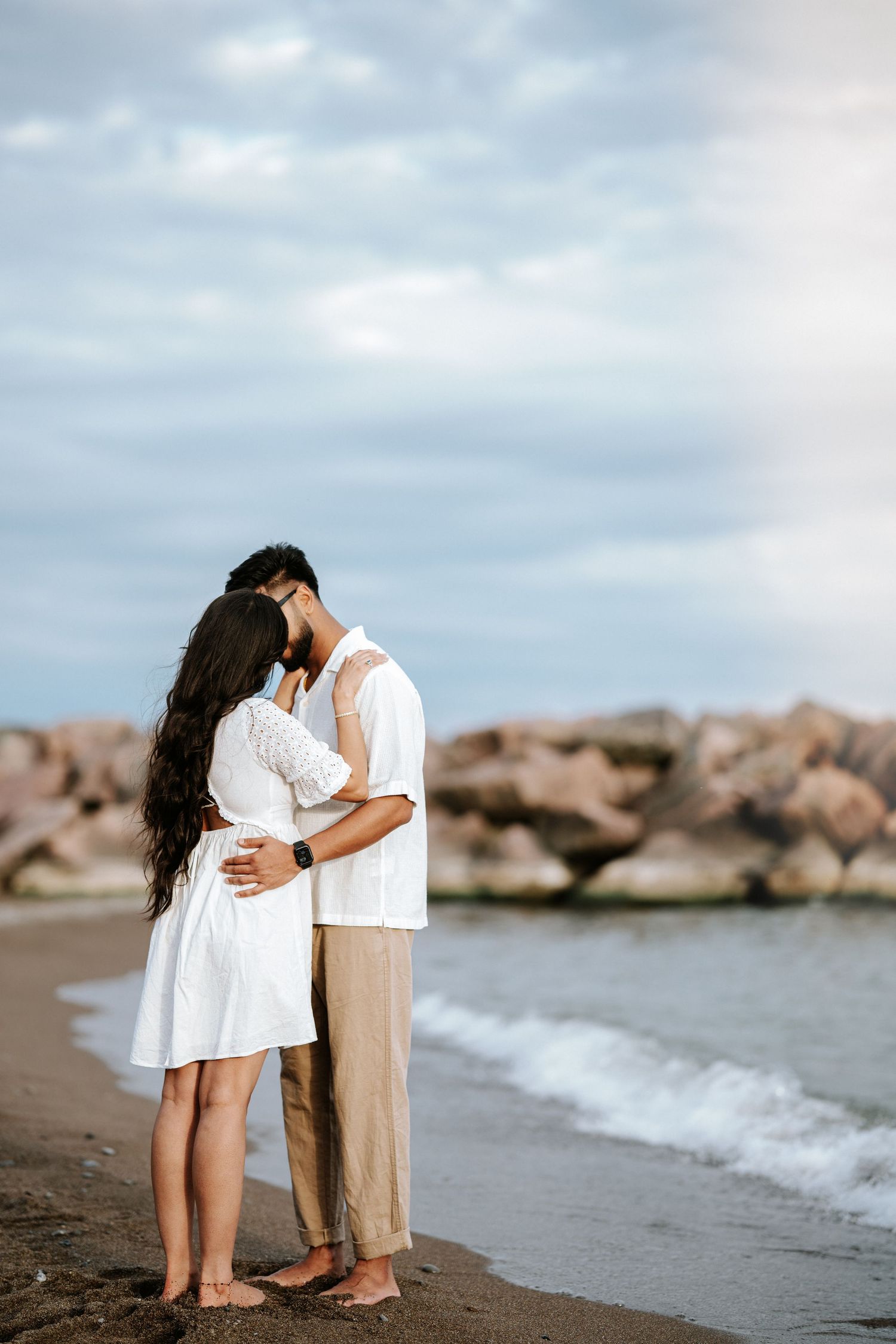 A tender moment shared on a beach with gentle waves and rocky shoreline.