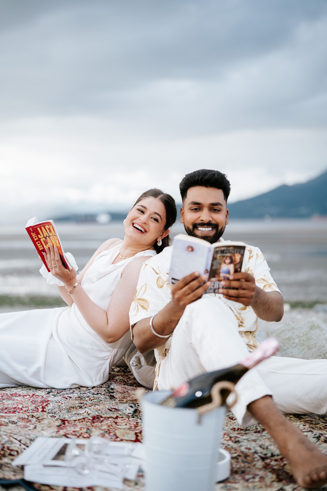 Smiling couple sitting on beach blanket, woman holding McDonald's cup and man showing phone, mountains behind.