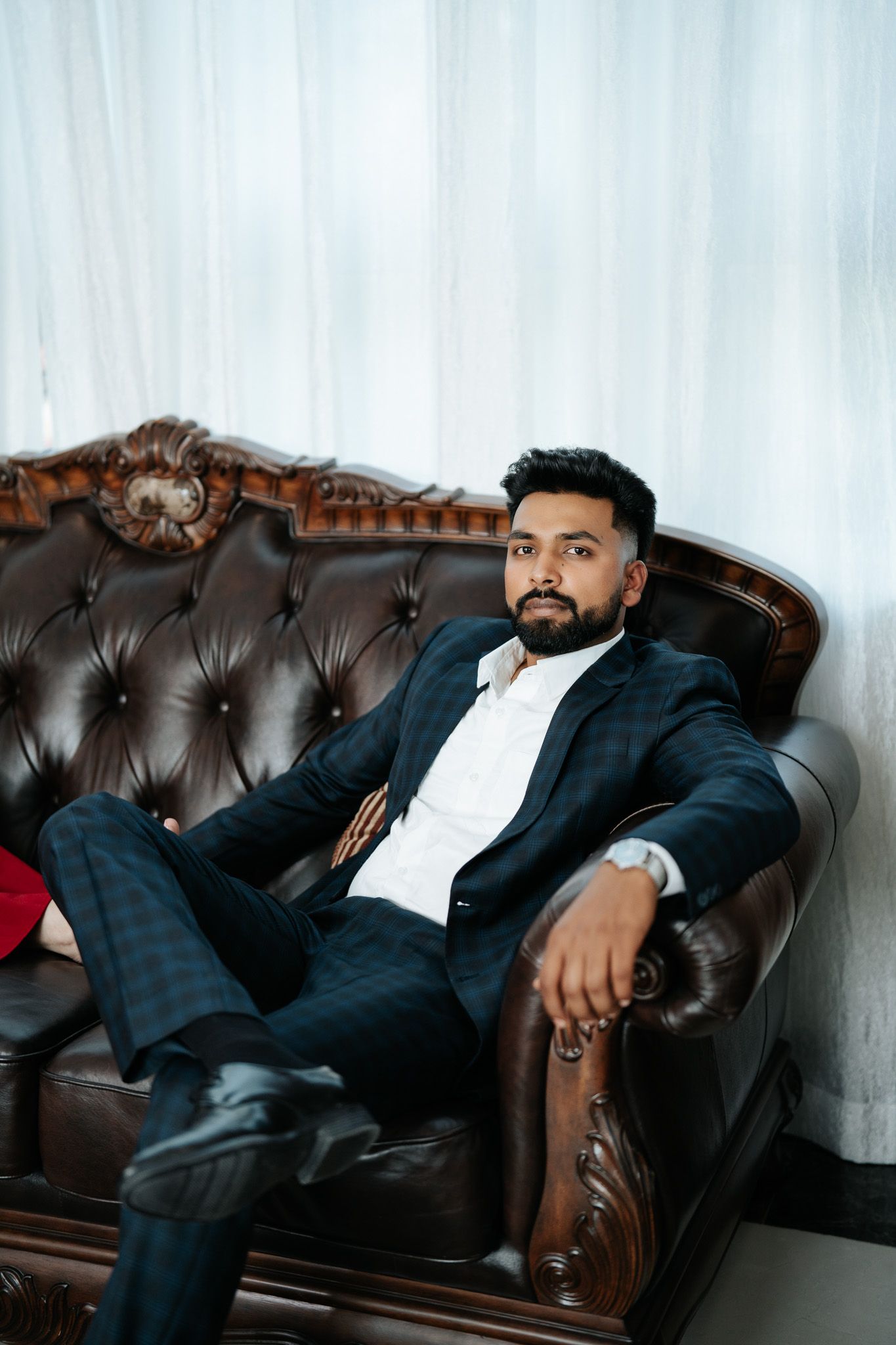 Stylish bearded man in dark suit relaxing confidently on tufted brown leather sofa.