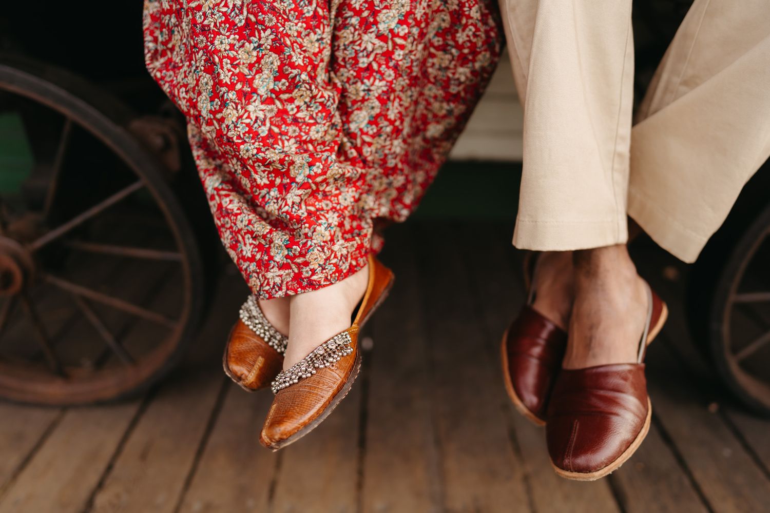 Close-up of a couple's feet in traditional Indian footwear, woman in red floral dress and man in tan trousers.
