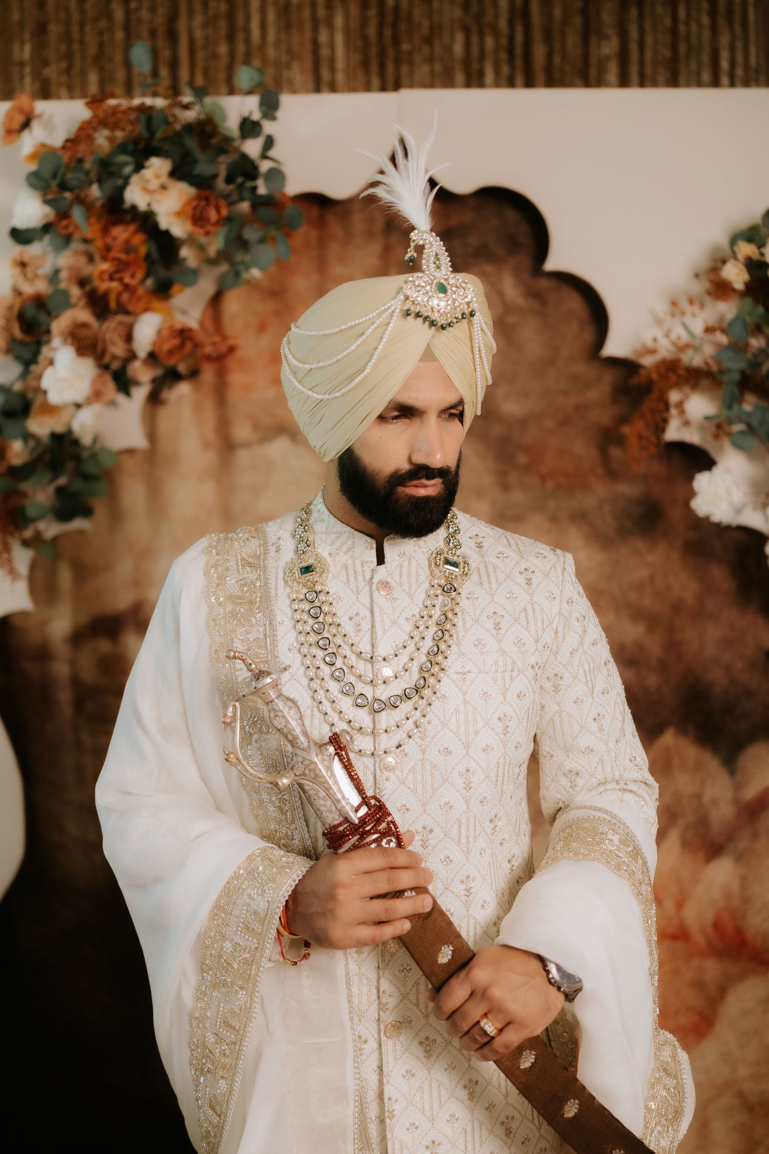 Sikh groom in white sherwani and turban holding a ceremonial sword at a wedding.