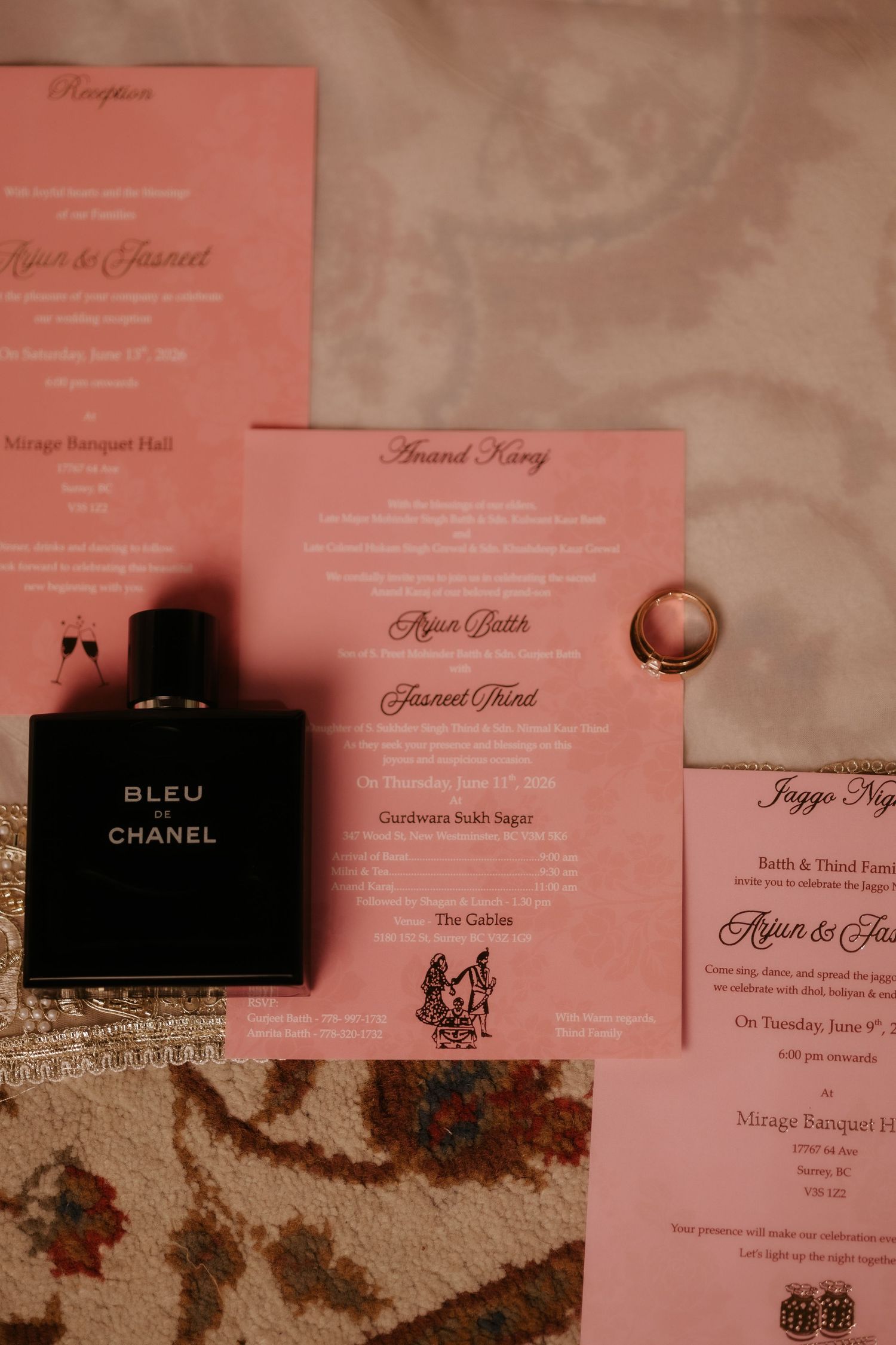 Chanel Bleu cologne beside pink wedding invitations for Arjun and Jasneet with a gold ring on a decorative surface.