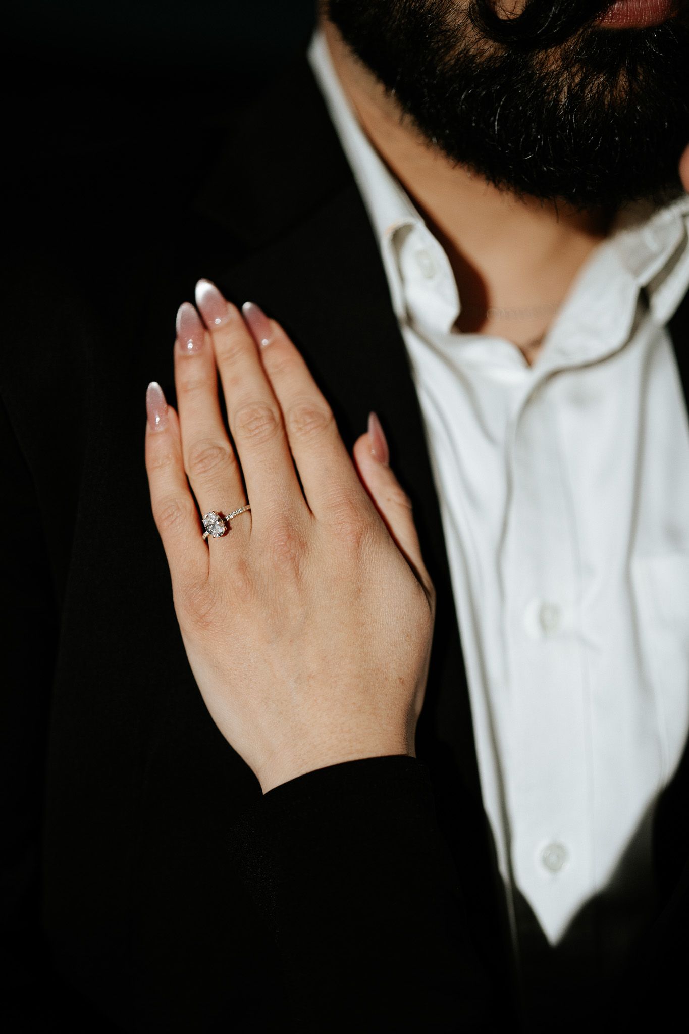 A close-up shot shows clasped hands with an engagement ring against a white shirt and dark background.