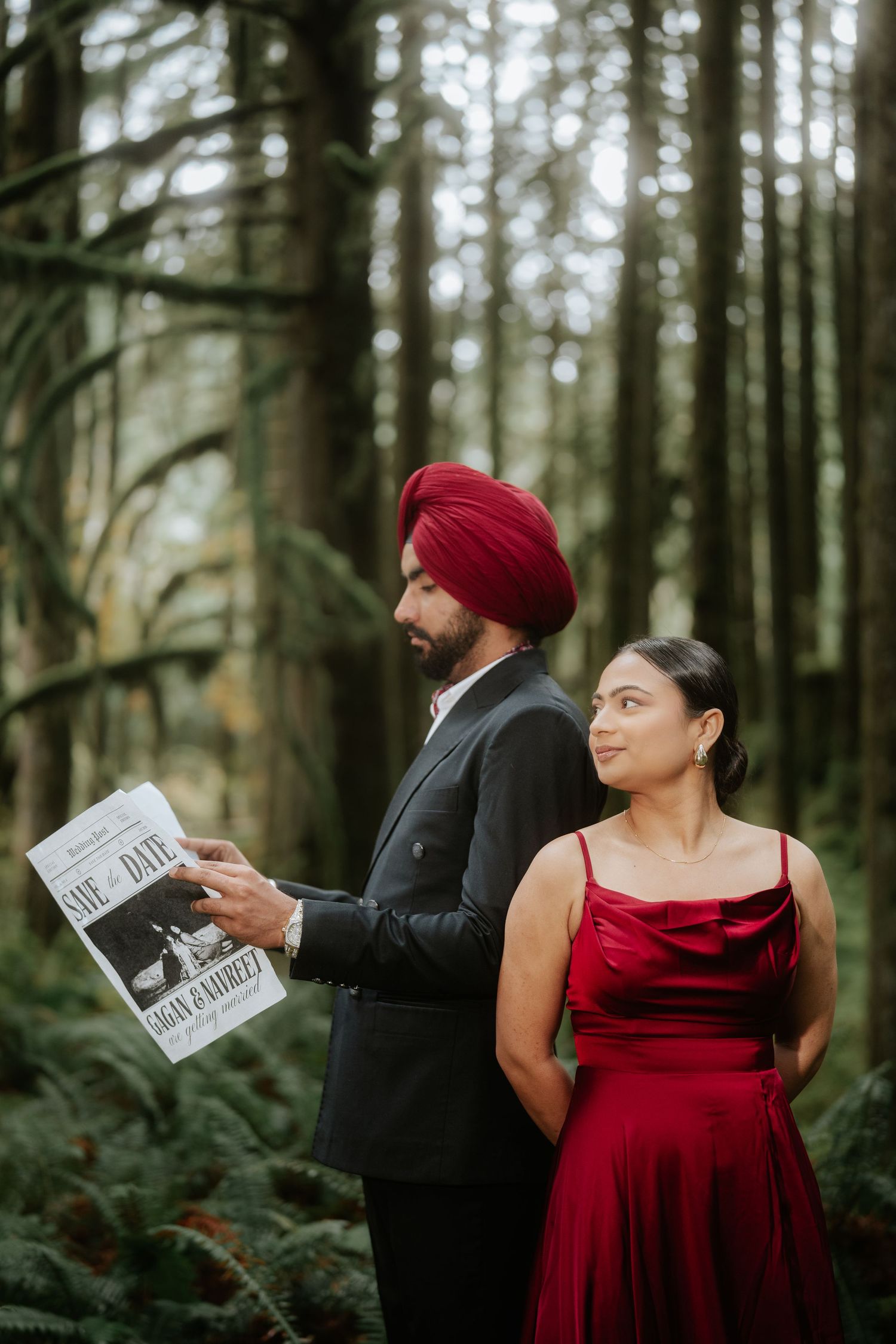 Couple in red and black formal attire poses with newspapers in evergreen forest.