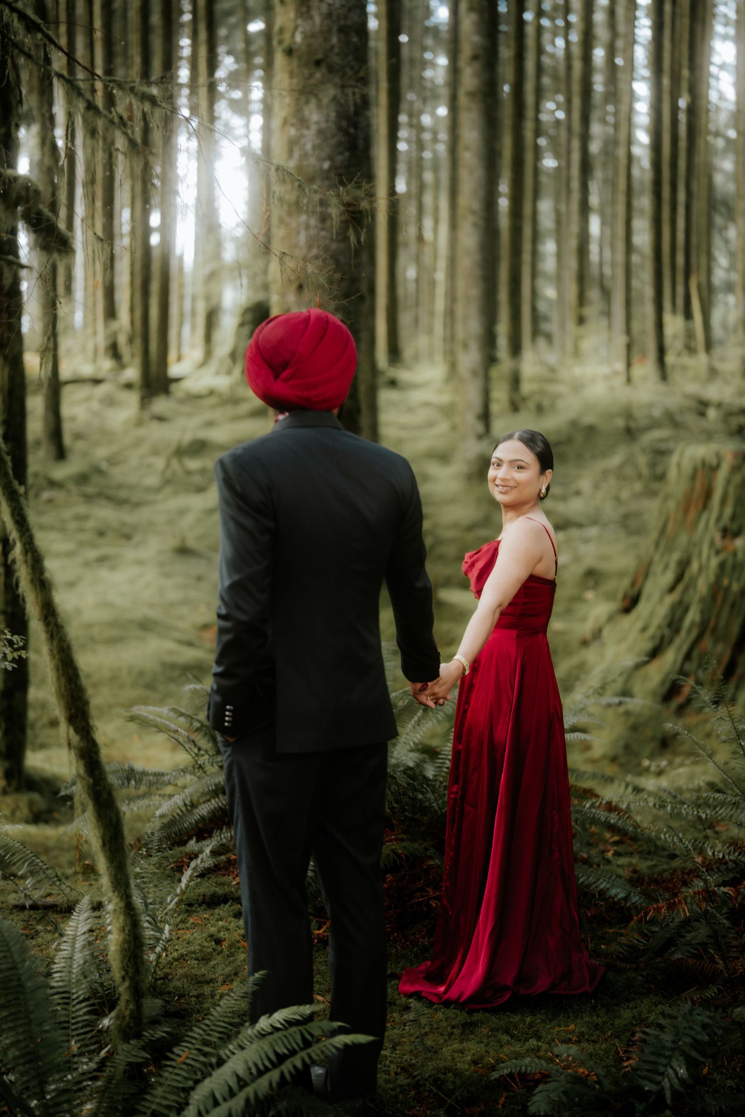 A couple in formal attire poses in a misty forest setting with tall trees and ferns.