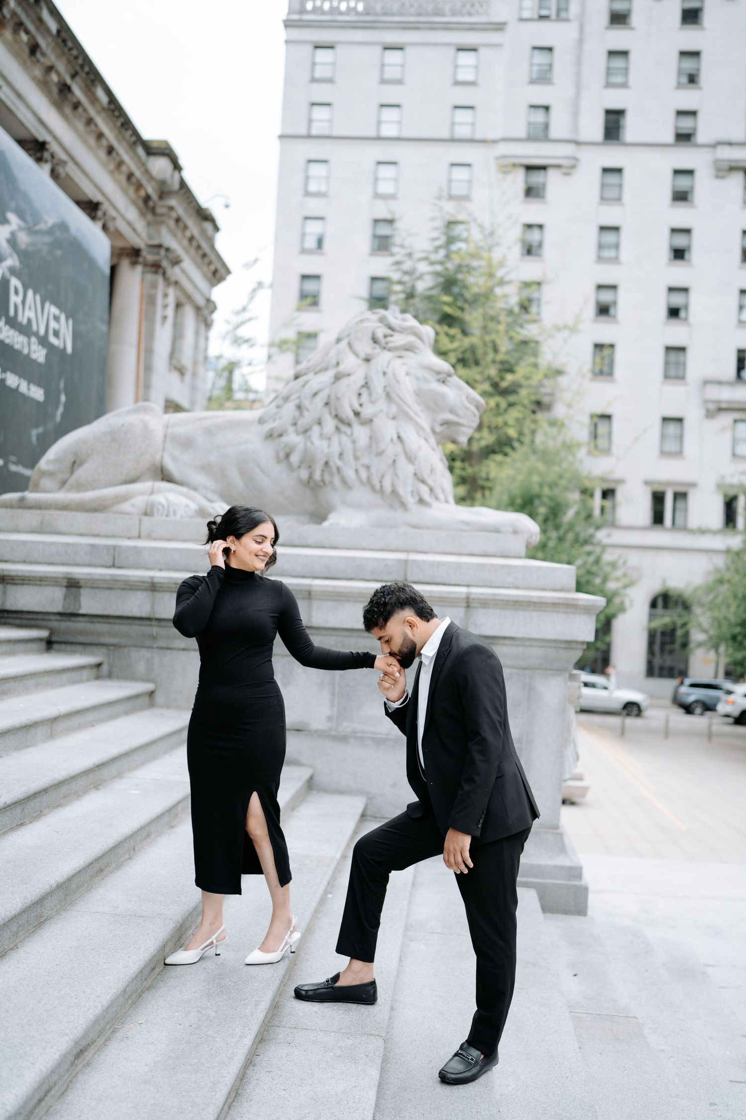 A playful couple poses together on wide marble steps near ornate stone sculptures.