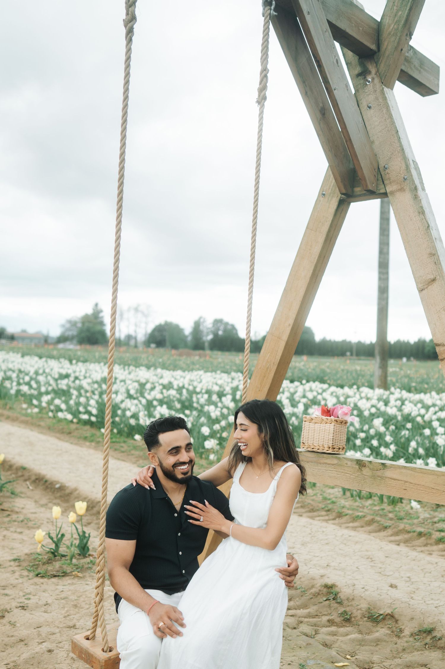 A couple shares a romantic moment on a swing in a blooming white flower field on a cloudy day.