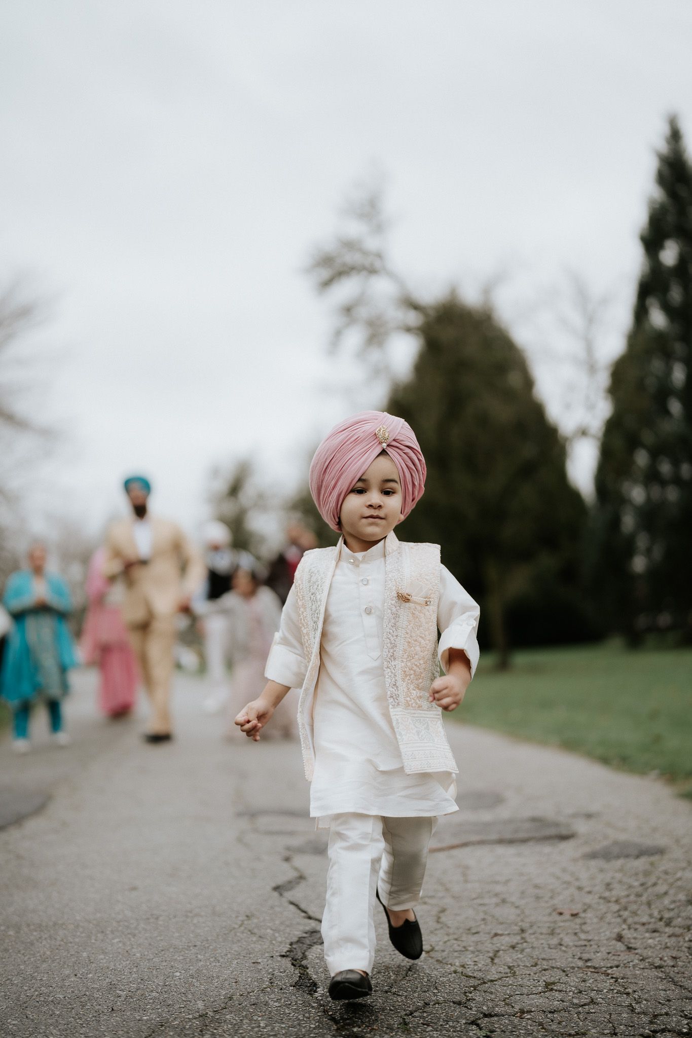 A small child in a white traditional outfit and pink turban runs joyfully on a path while others walk behind.