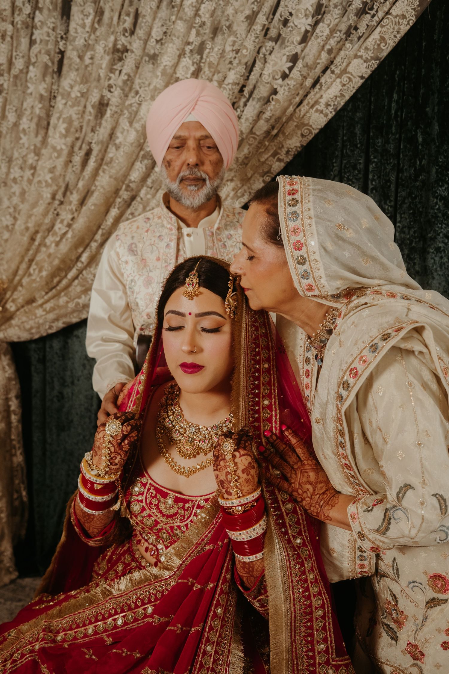Sikh bride in red lehenga with gold jewelry receives blessings from parents in traditional attire at wedding ceremony.