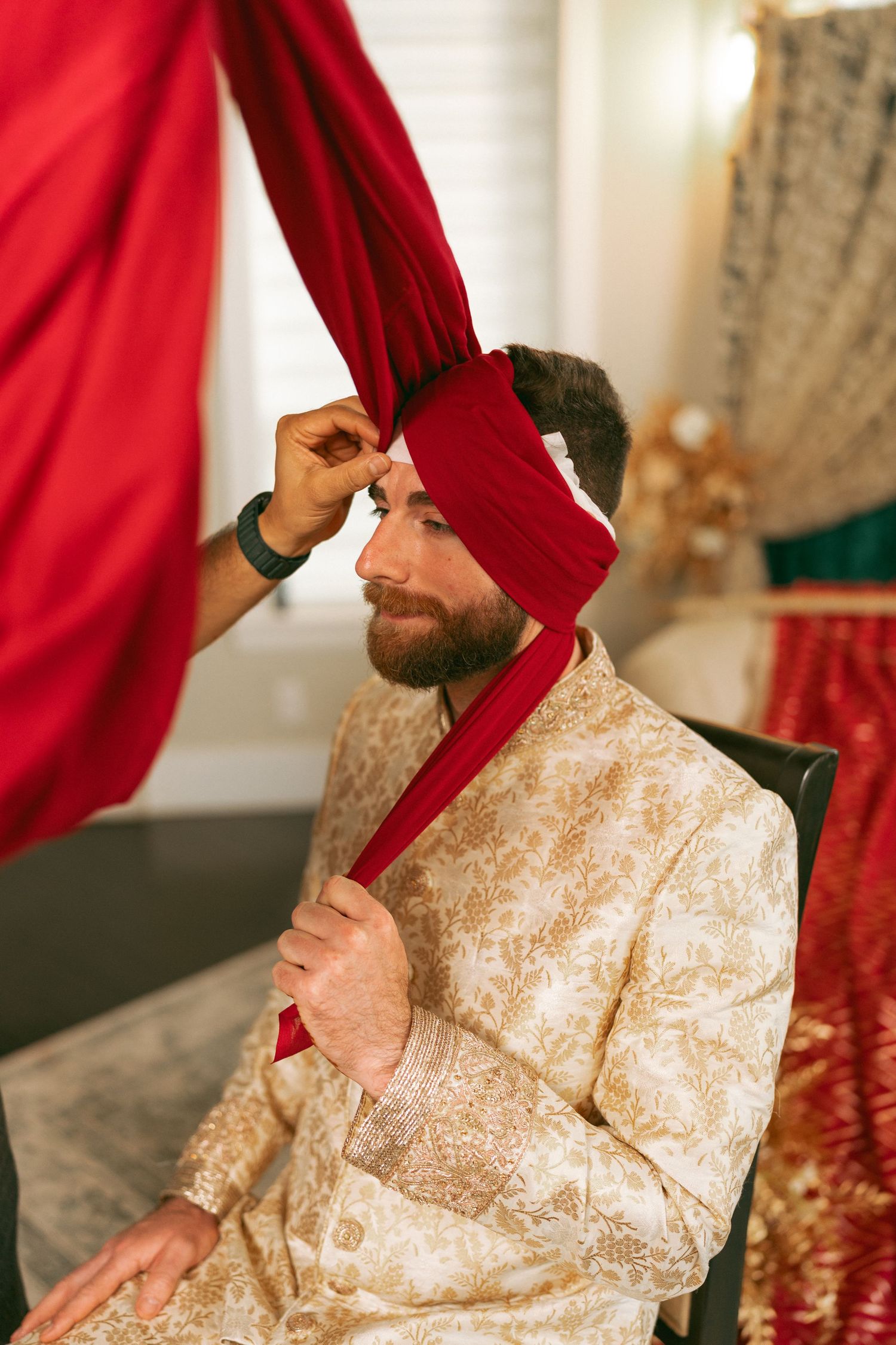 Groom in gold sherwani having red turban tied during Indian wedding preparation ceremony.