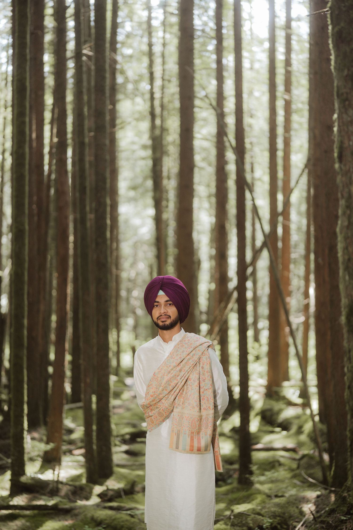Sikh man in white kurta and burgundy turban with paisley shawl standing in sunlit forest.