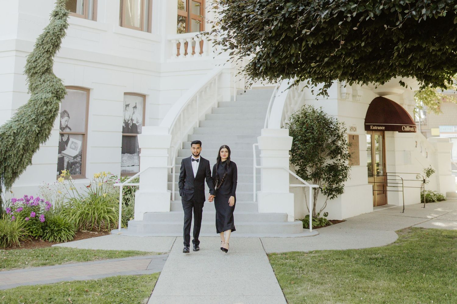 Stylish couple in black formal wear walking hand in hand outside a white classical architecture building.