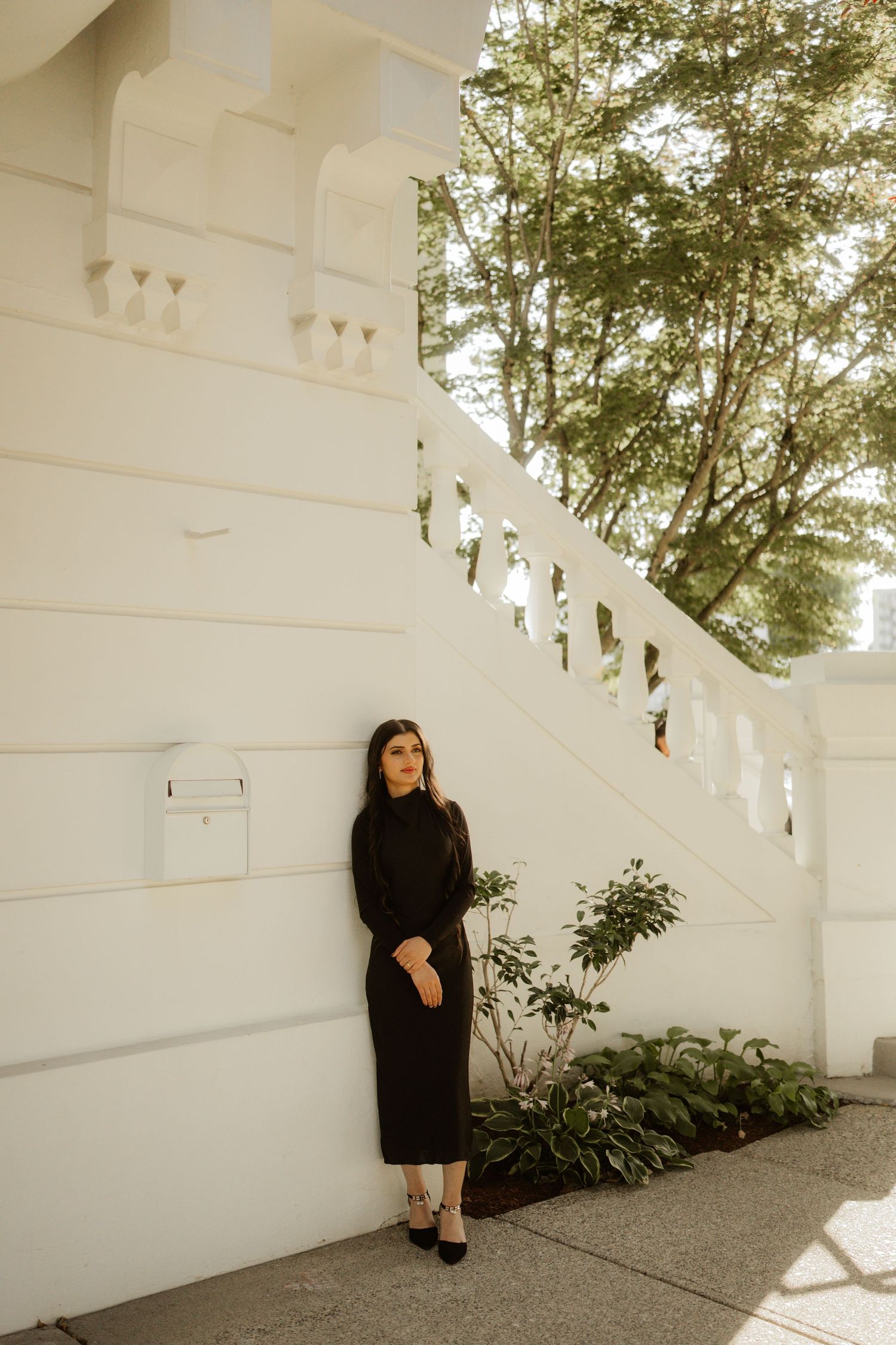 Woman in black dress leaning against white wall with balustrade and green trees.