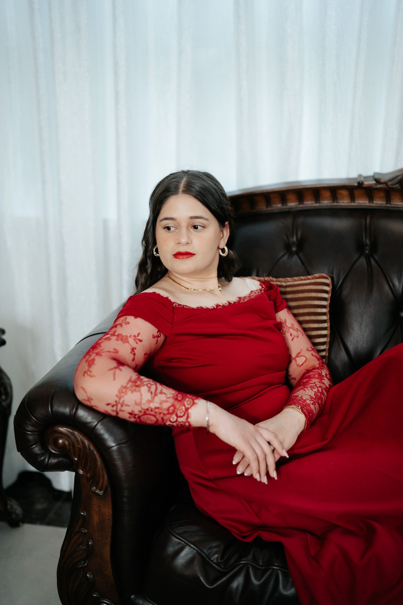 Woman in red lace-sleeve gown seated on a tufted leather sofa with a sophisticated expression.