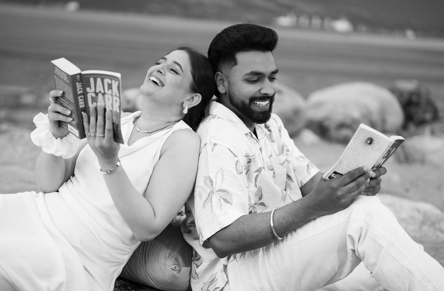 Black and white photo of a laughing couple sitting back to back outdoors, both holding books and smiling.