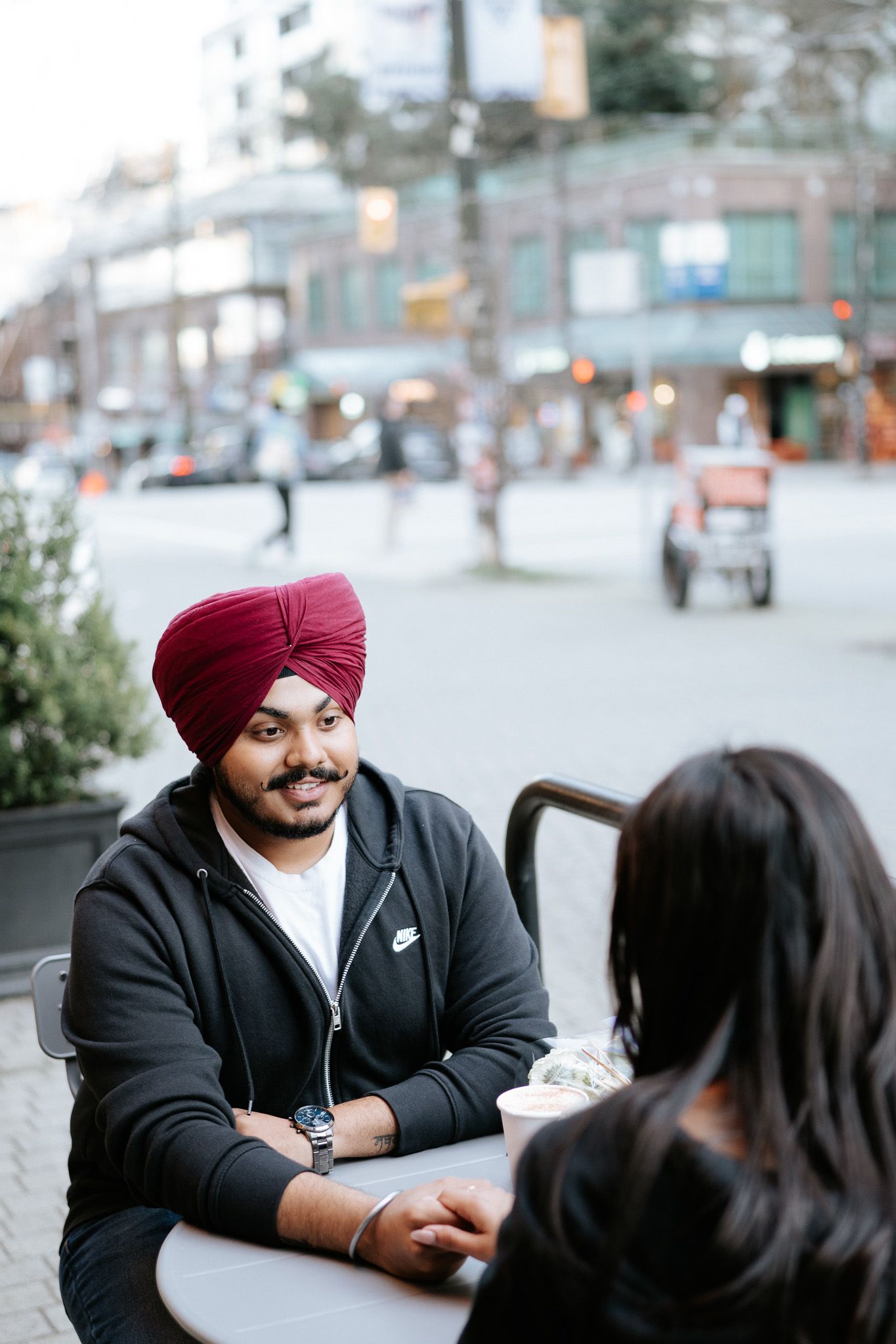 A person wearing a red turban and black jacket smiles while sitting at an outdoor table during a conversation.