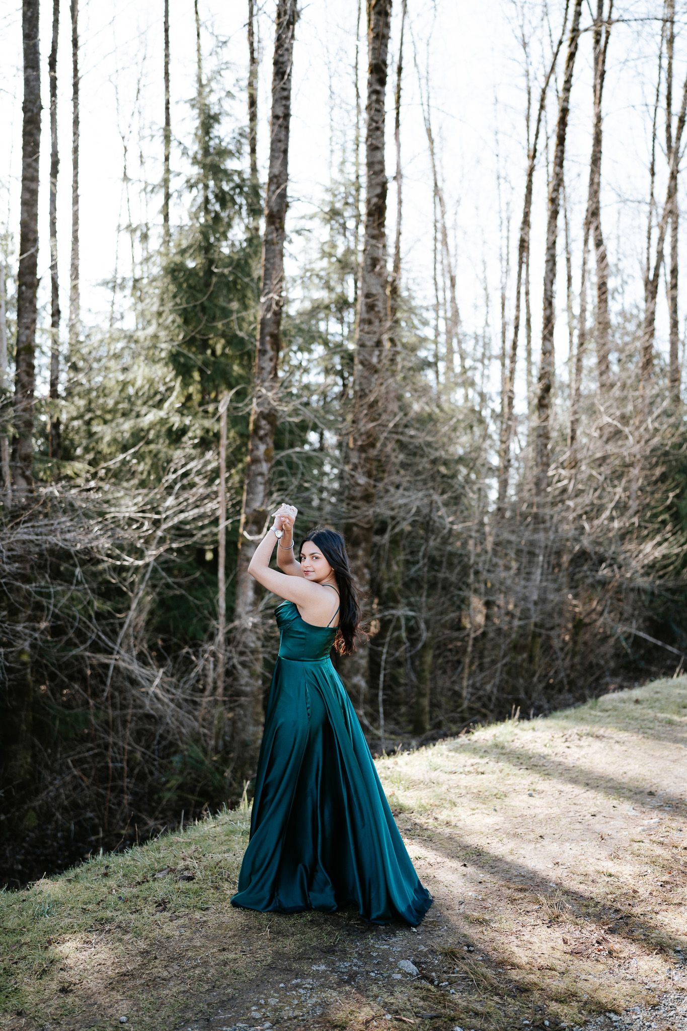A person in an elegant emerald green gown poses gracefully along a sunlit forest path in winter.