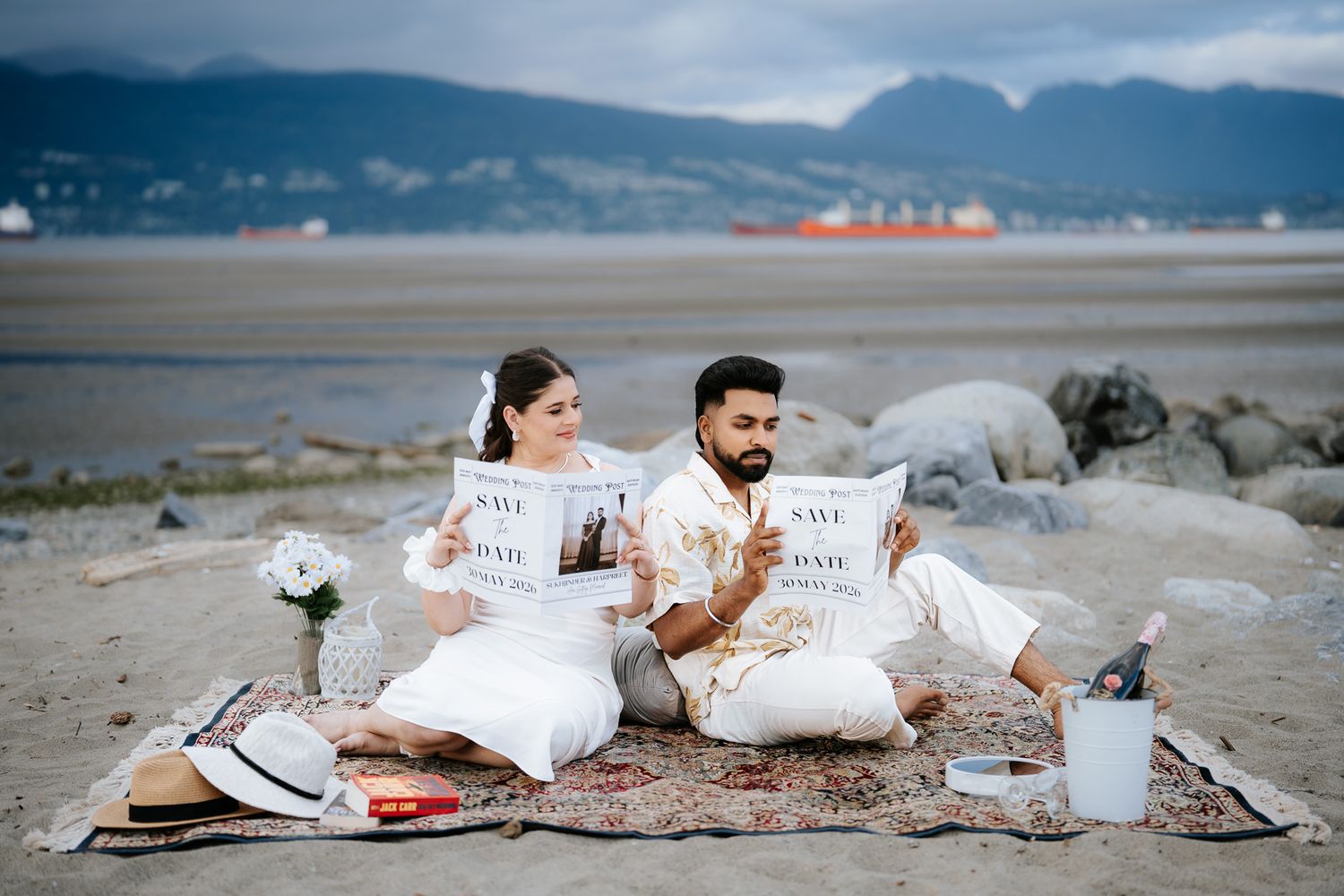 Engaged couple seated on blanket at sandy beach displaying Save the Date cards with scenic mountain backdrop.