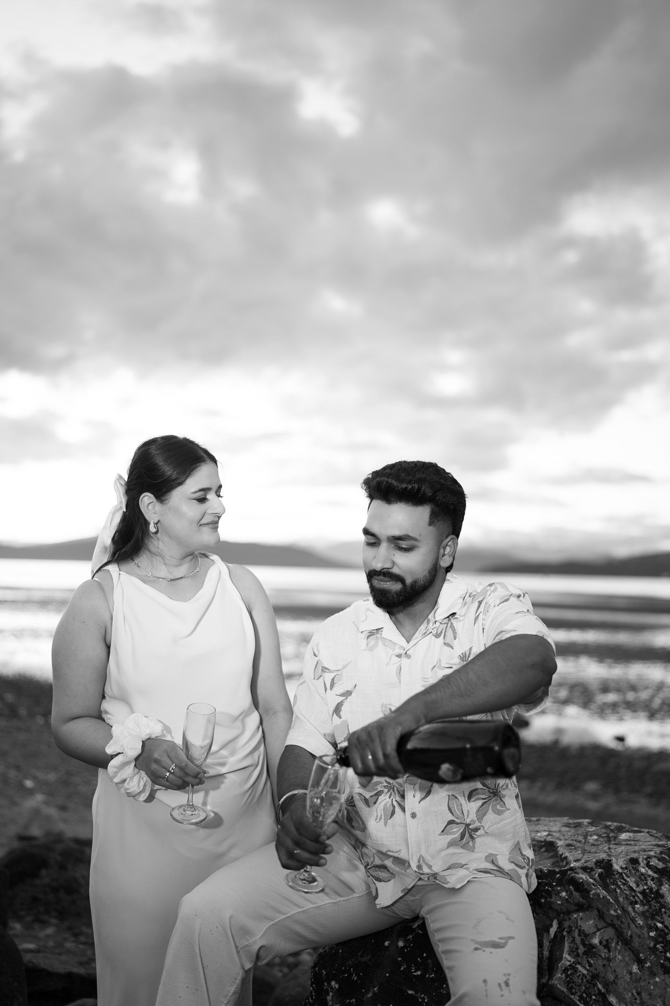 Black and white photo of a couple at the beach, man pouring champagne into glasses held by woman at sunset.