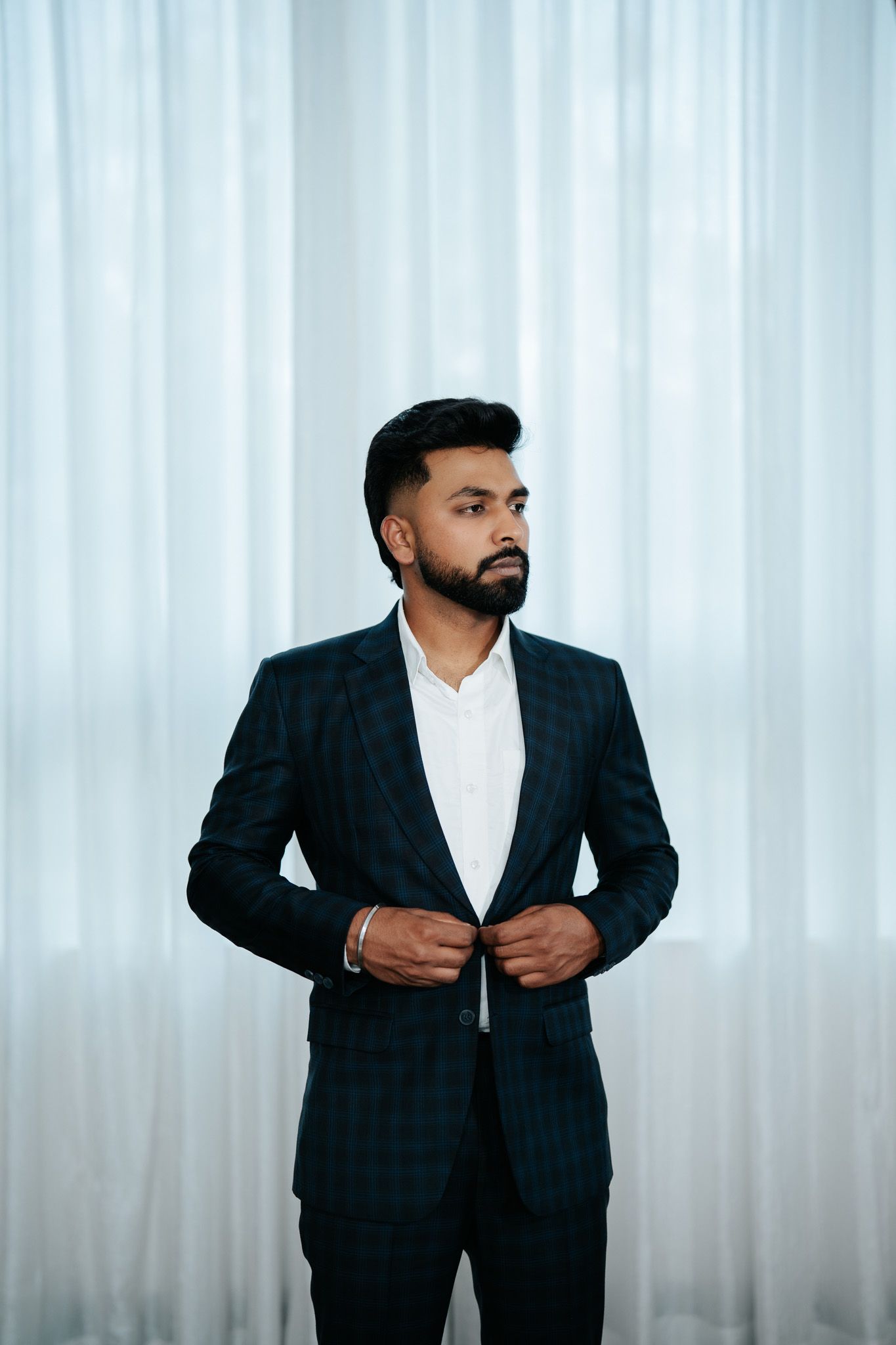 Confident bearded man in dark plaid suit buttoning jacket while standing before sheer white curtains.