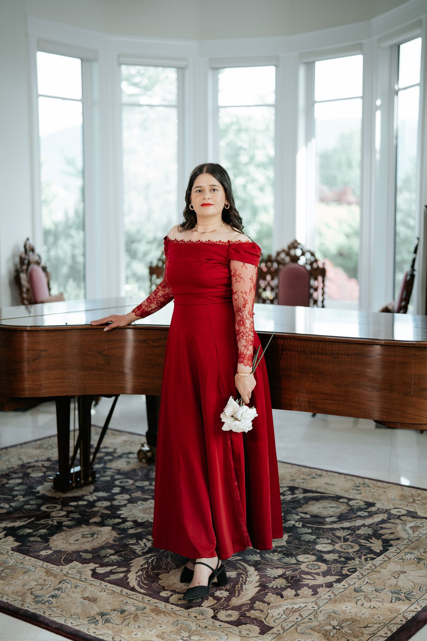 Woman in elegant red off-shoulder lace gown holding white flowers beside a grand piano.