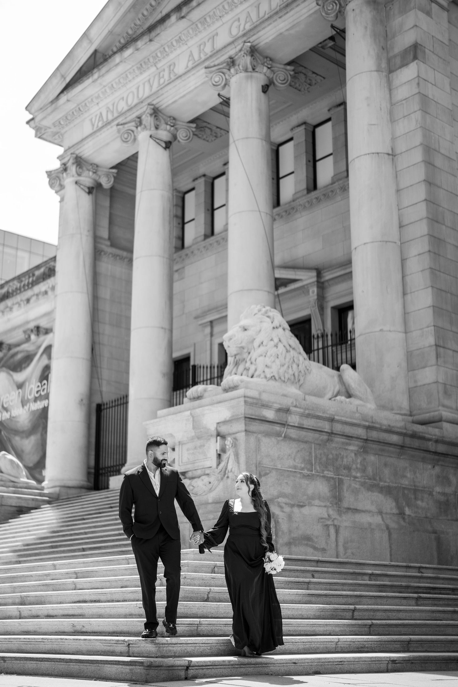 Couple holding hands on steps of Vancouver Art Gallery with lion statue, black and white wedding photo.