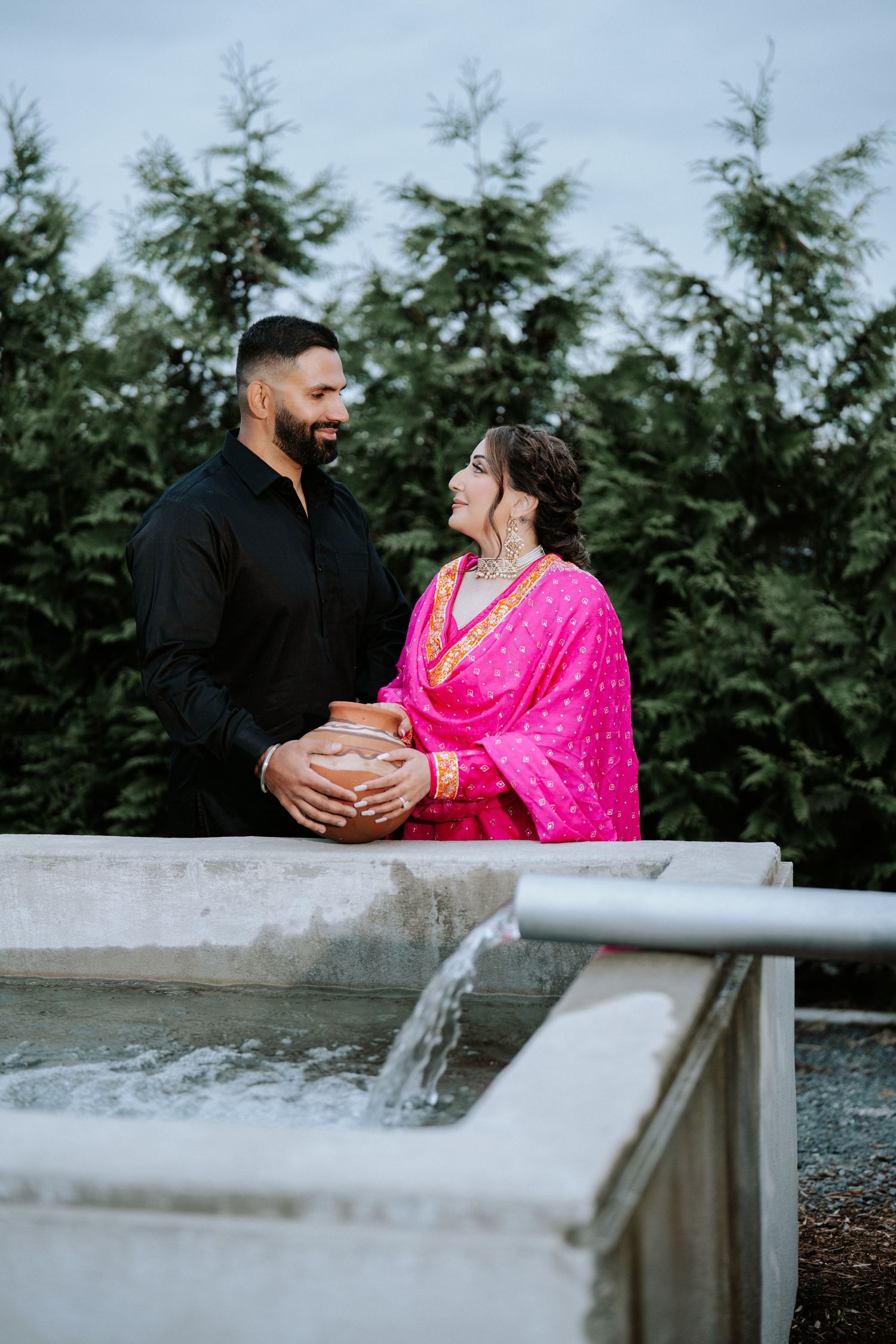 A couple in traditional South Asian attire poses lovingly by a fountain, surrounded by lush green trees.