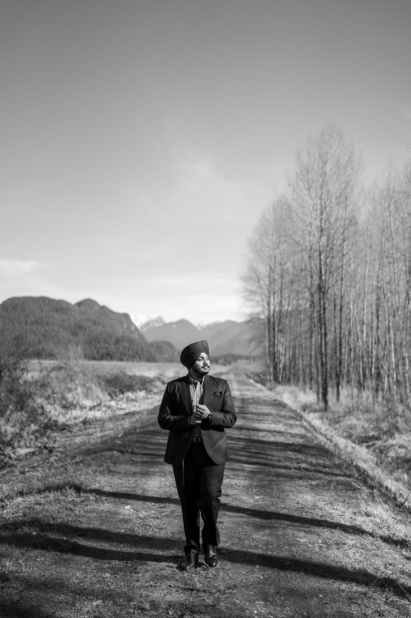 Black and white photo of a person standing on a rural mountain road surrounded by bare trees and distant peaks.