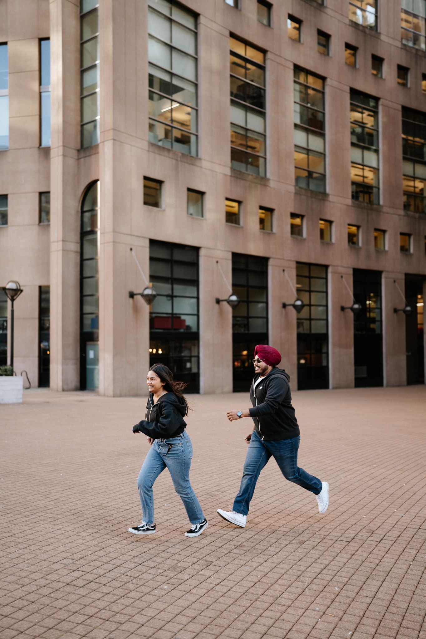 Two people perform a playful dance move sequence in front of a modern office building with stone walls and glass windows.