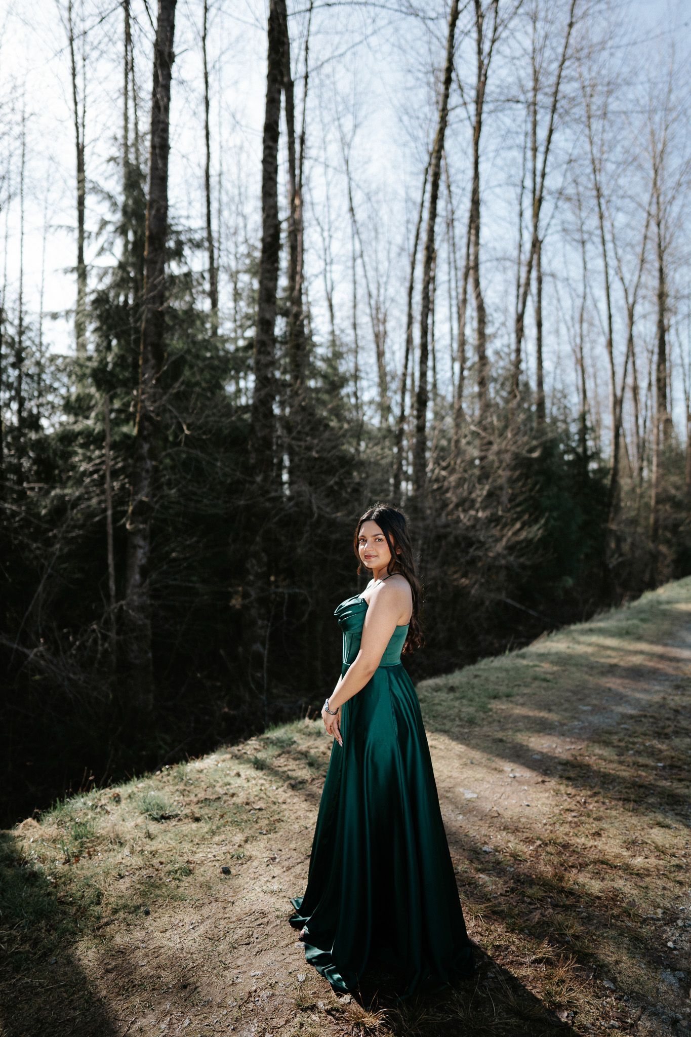 A person wearing a flowing dark green dress poses on a forest path with tall bare trees in winter.