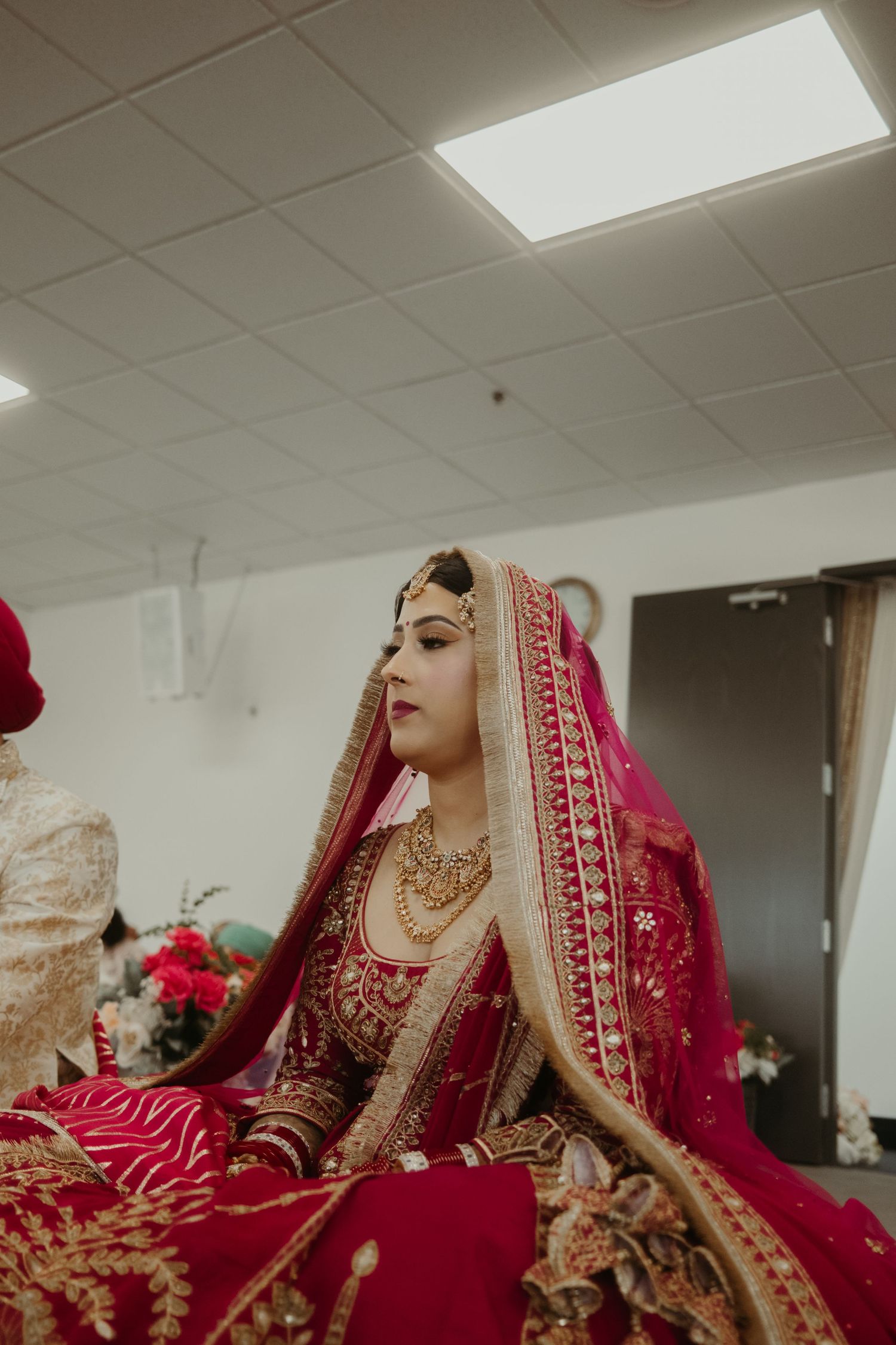A bride in a red and gold embroidered lehenga with matching dupatta and traditional gold jewelry sits gracefully indoors.