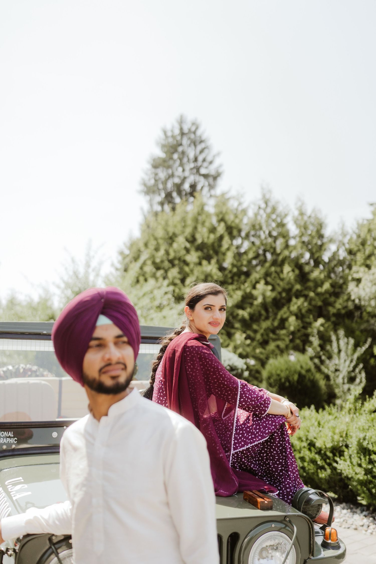 Sikh couple in purple and white traditional wear pose near a vintage green jeep outdoors.