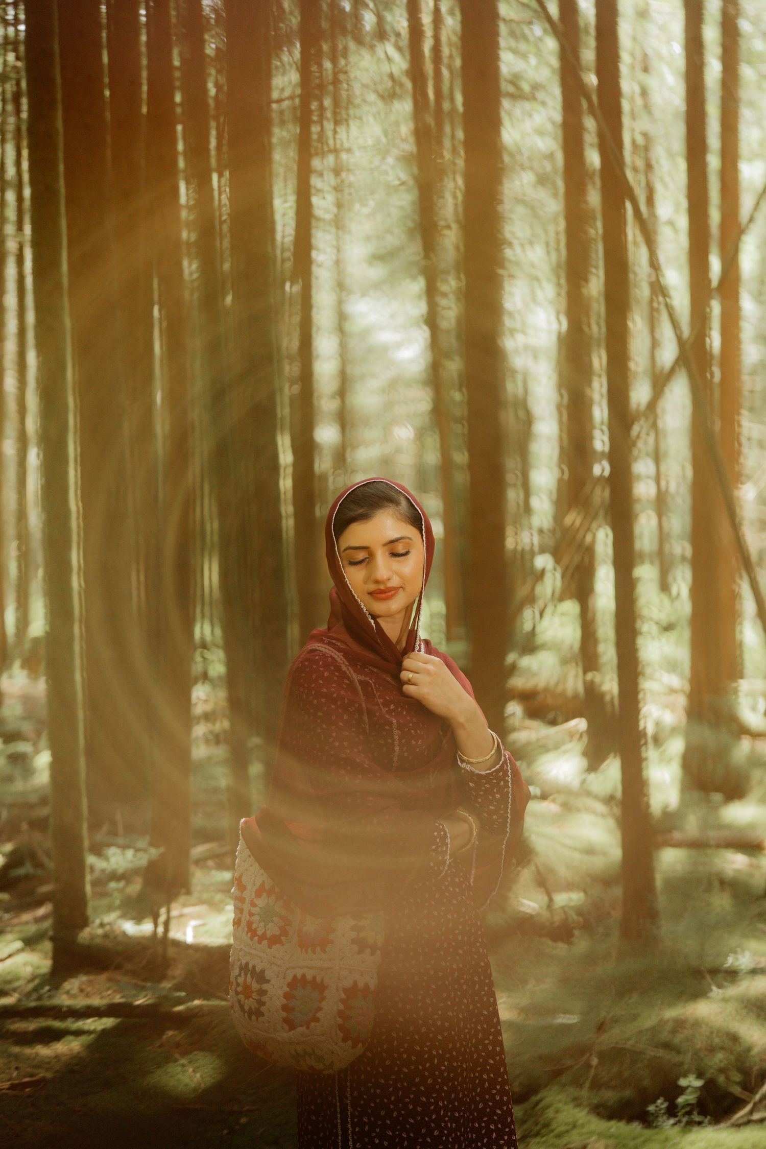 A woman in traditional South Asian attire and head covering stands peacefully in a sunlit forest.