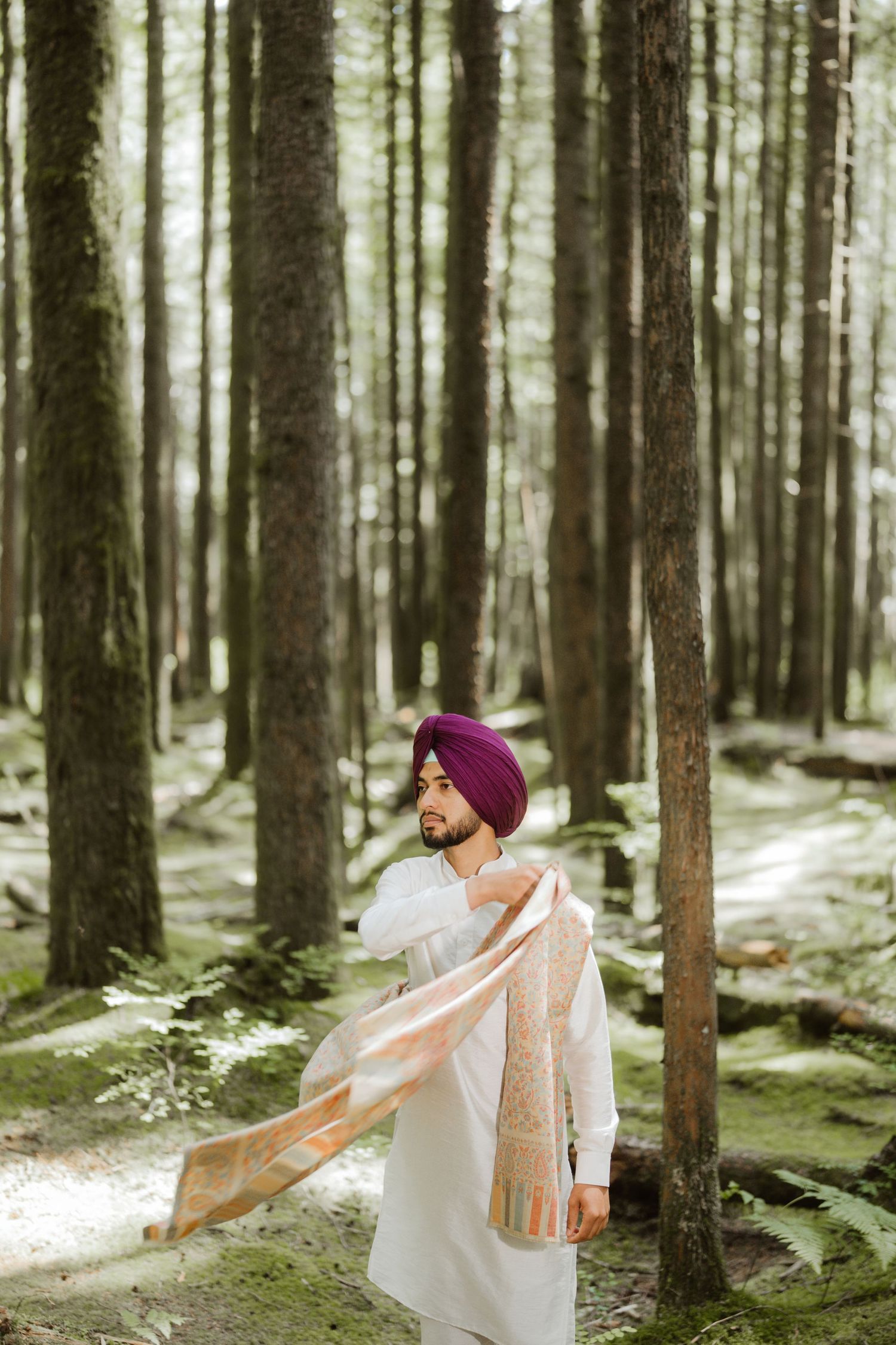 A Sikh man in a purple turban and white kurta drapes a decorative shawl in a serene forest setting.