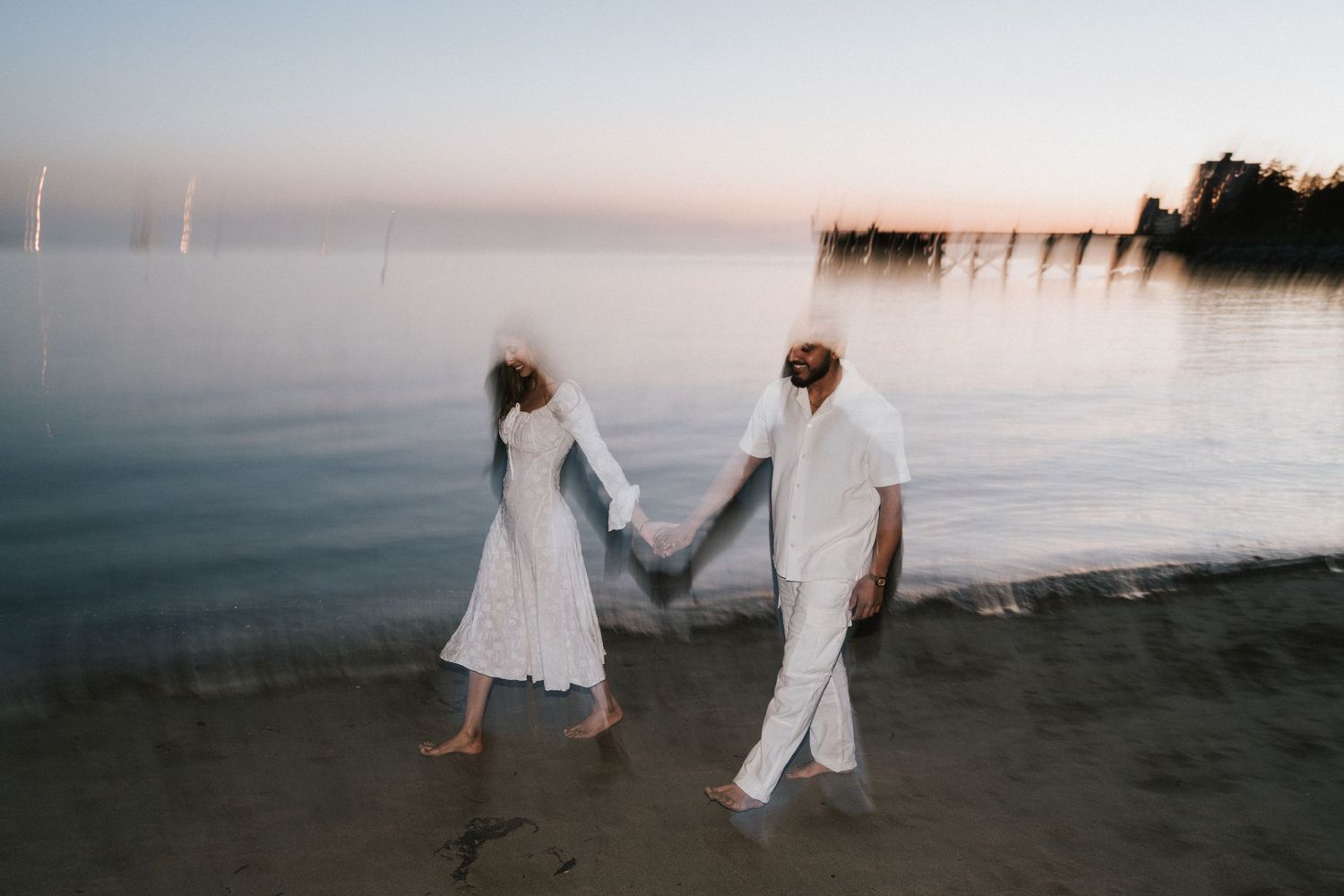 A couple in white outfits holds hands while walking along a misty beach at sunset.