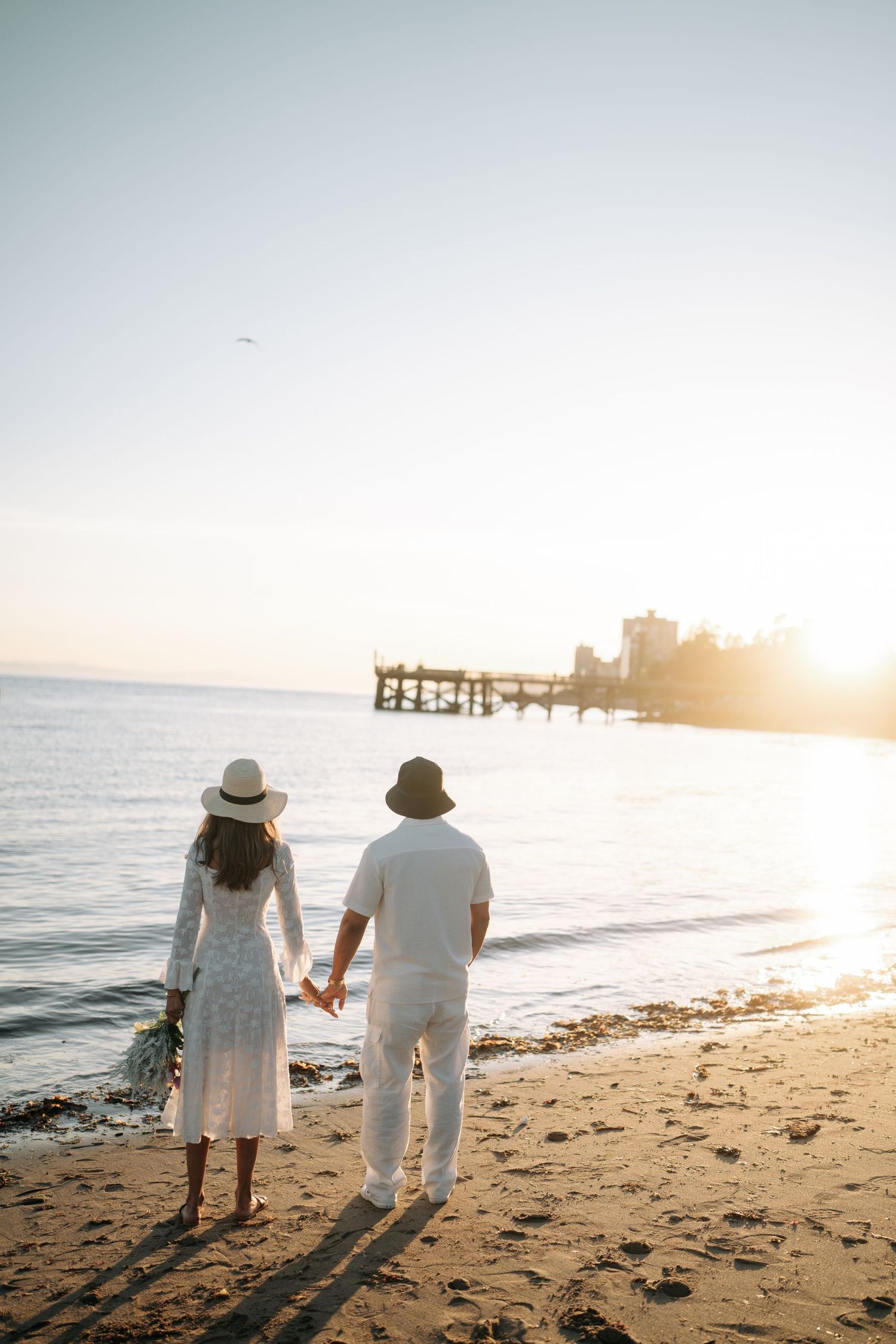 Couple in white outfits walking hand in hand along a sunset beach with a wooden pier in the distance.