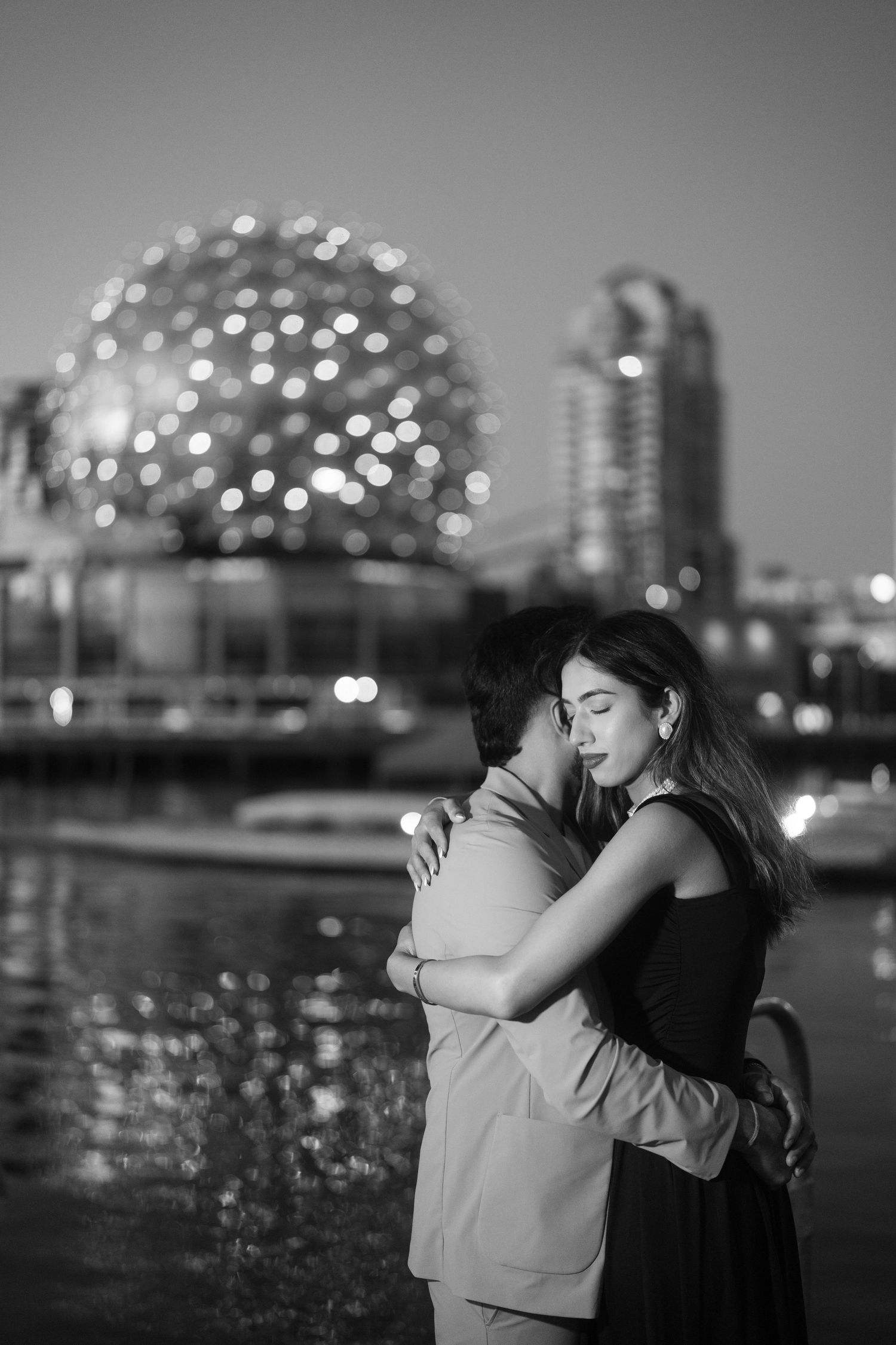 A romantic black and white photo series of a couple embracing by the waterfront with city lights twinkling in the background.