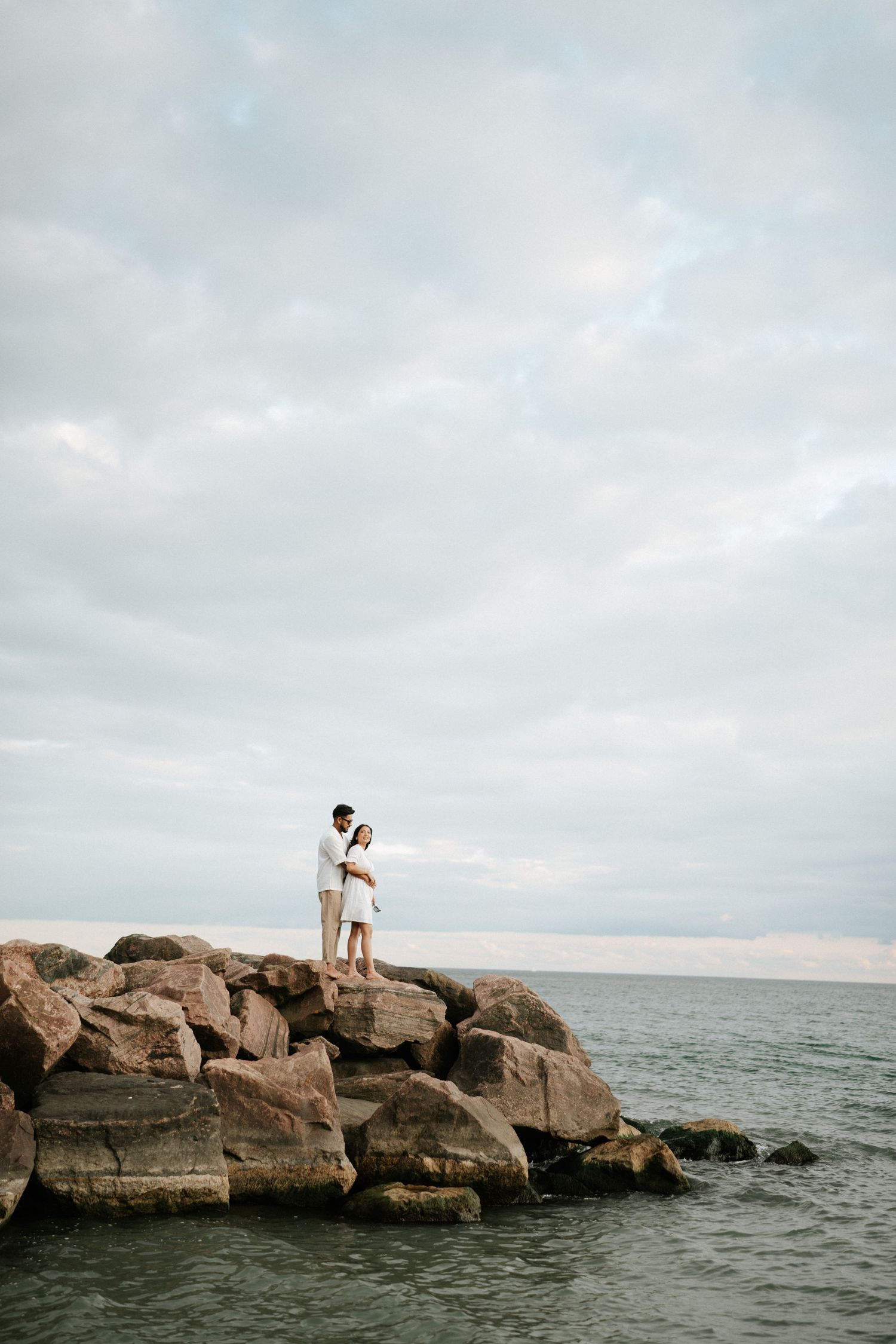 Two people stand on rocky ocean outcrop under cloudy skies.