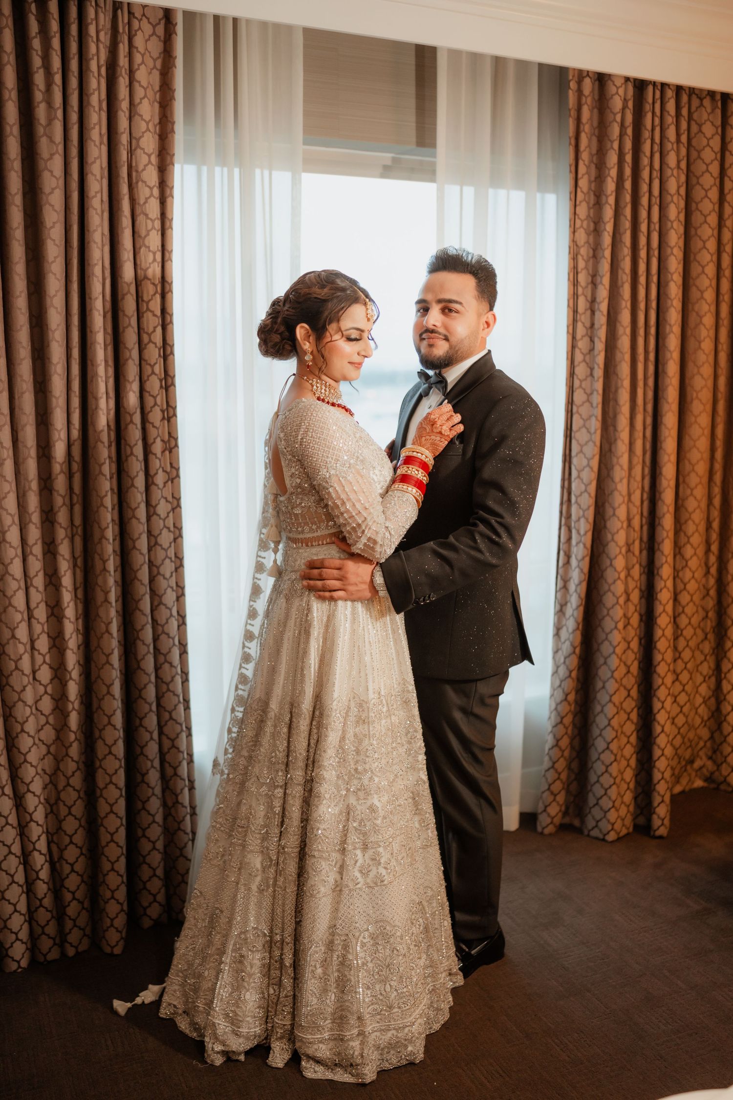 A couple in traditional wedding attire share a romantic dance near window with brown curtains.