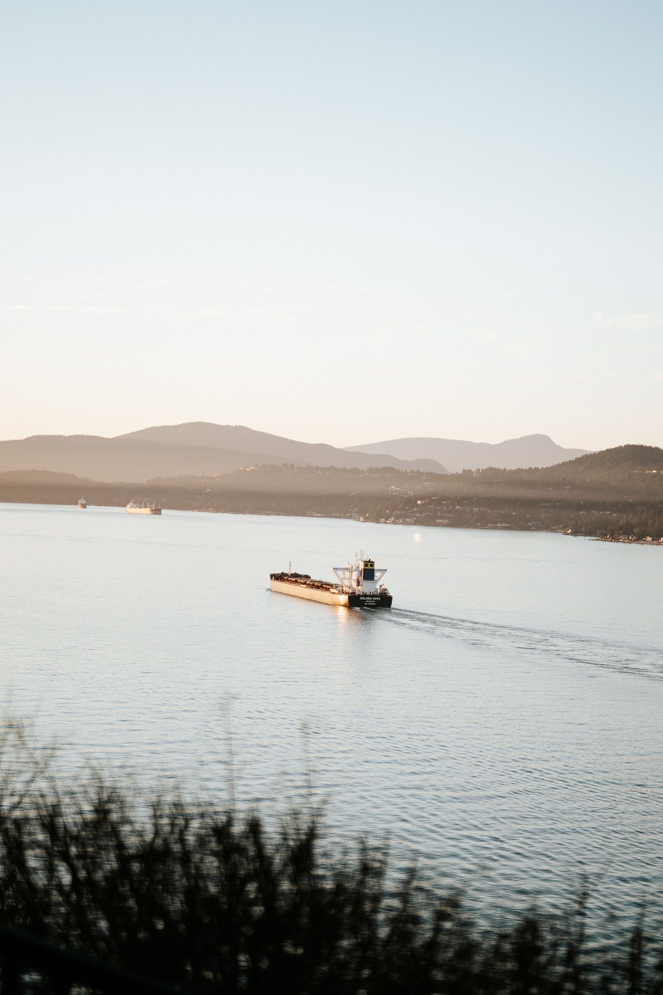 Cargo ship sailing across calm waters with distant mountains and hills visible in the background during sunset.