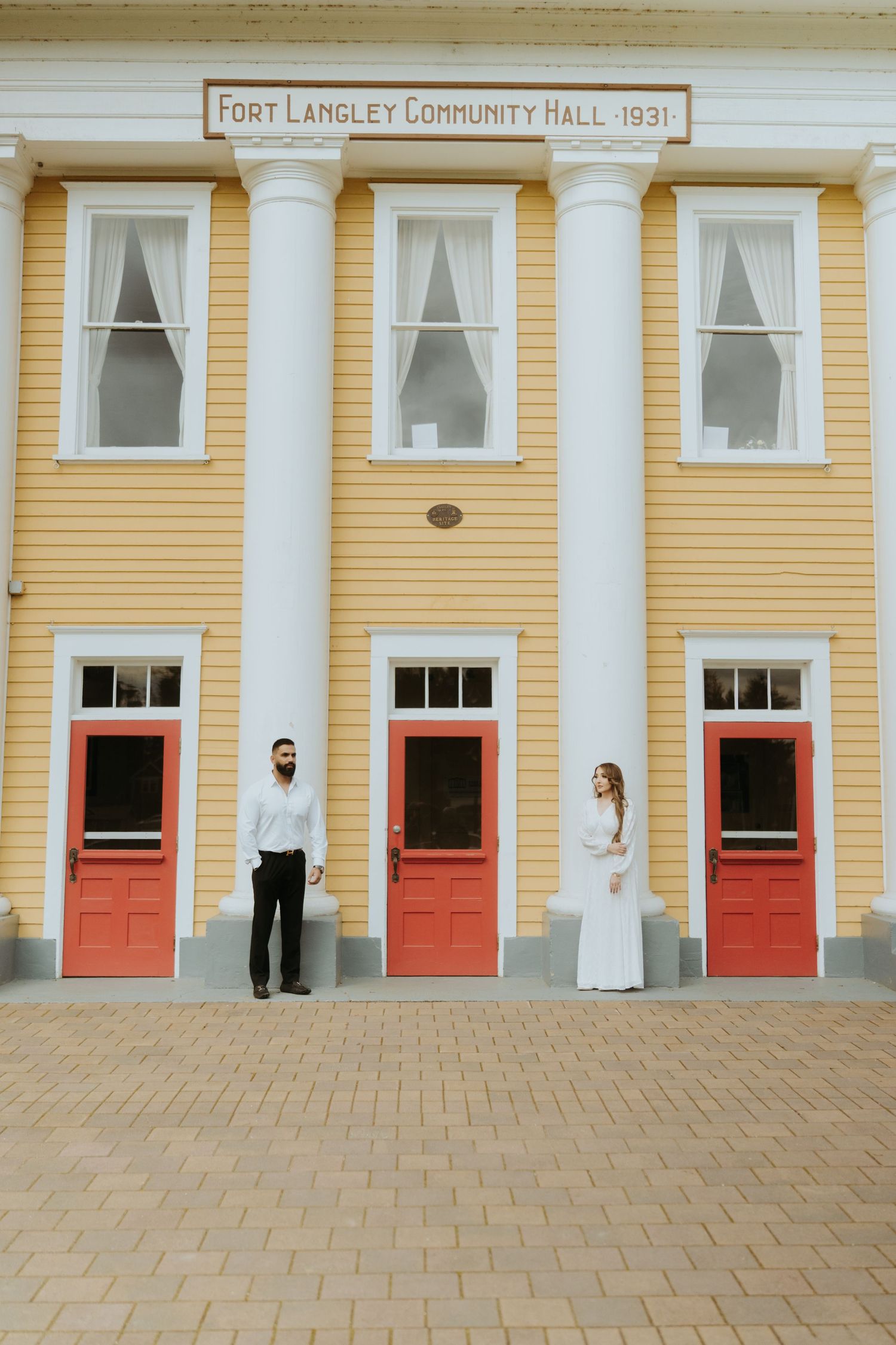 A couple poses in front of the Fort Langley Community Hall, a yellow building with red doors.